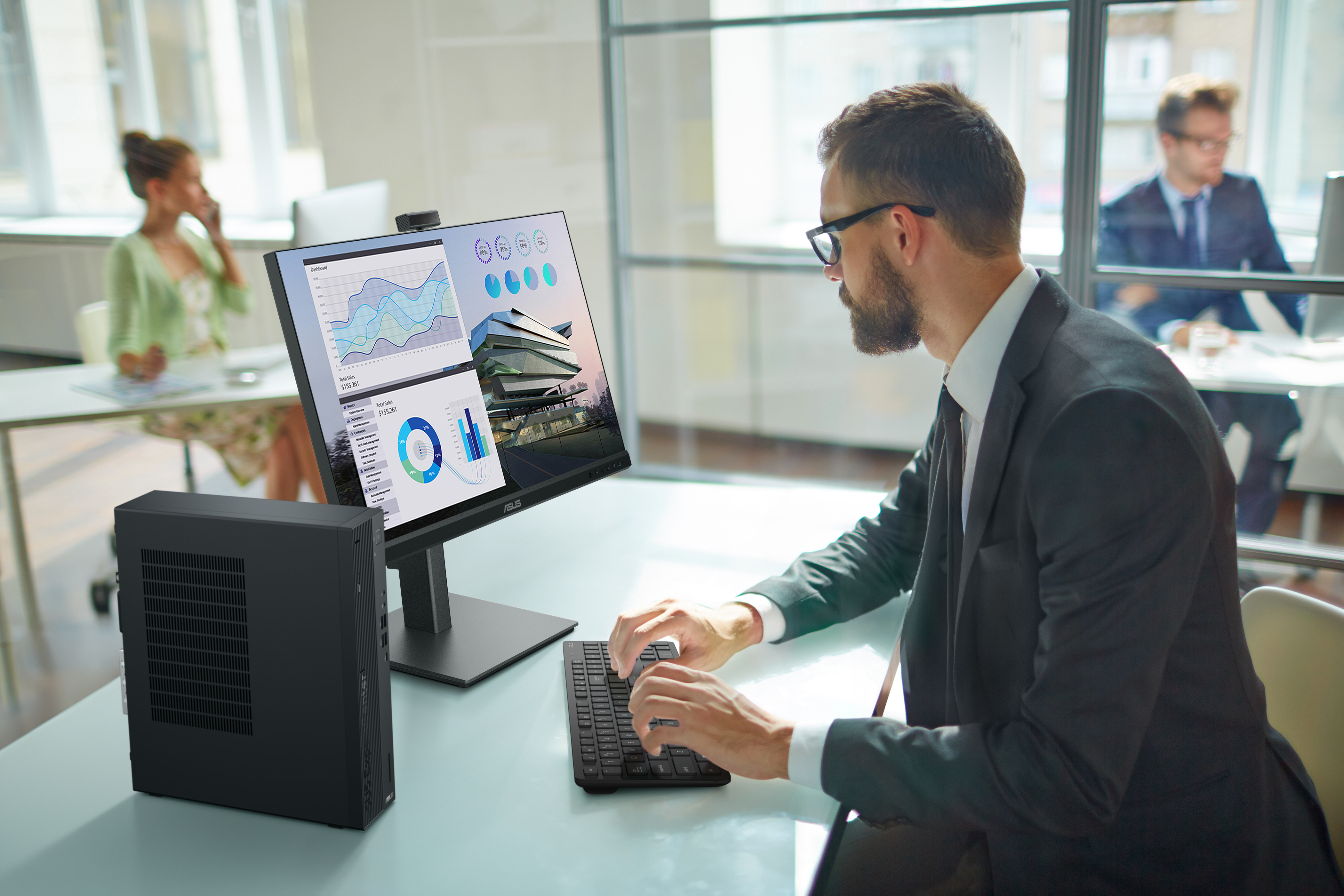 A man is working on a computer with charts on the screen. A black computer is on the left.