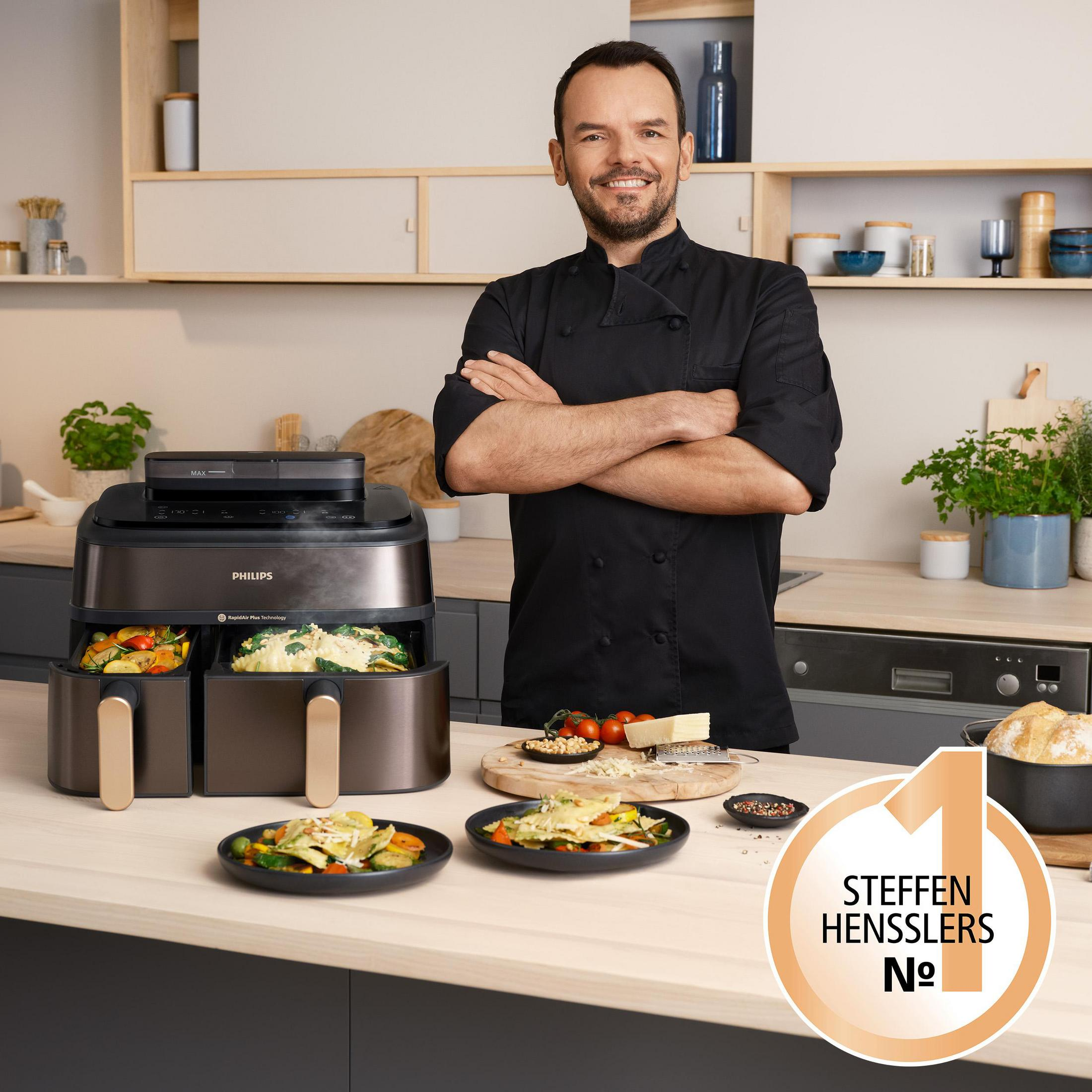 Szef kuchni w kuchni stoi obok czarnej frytownicy z jedzeniem i talerzami na blacie.