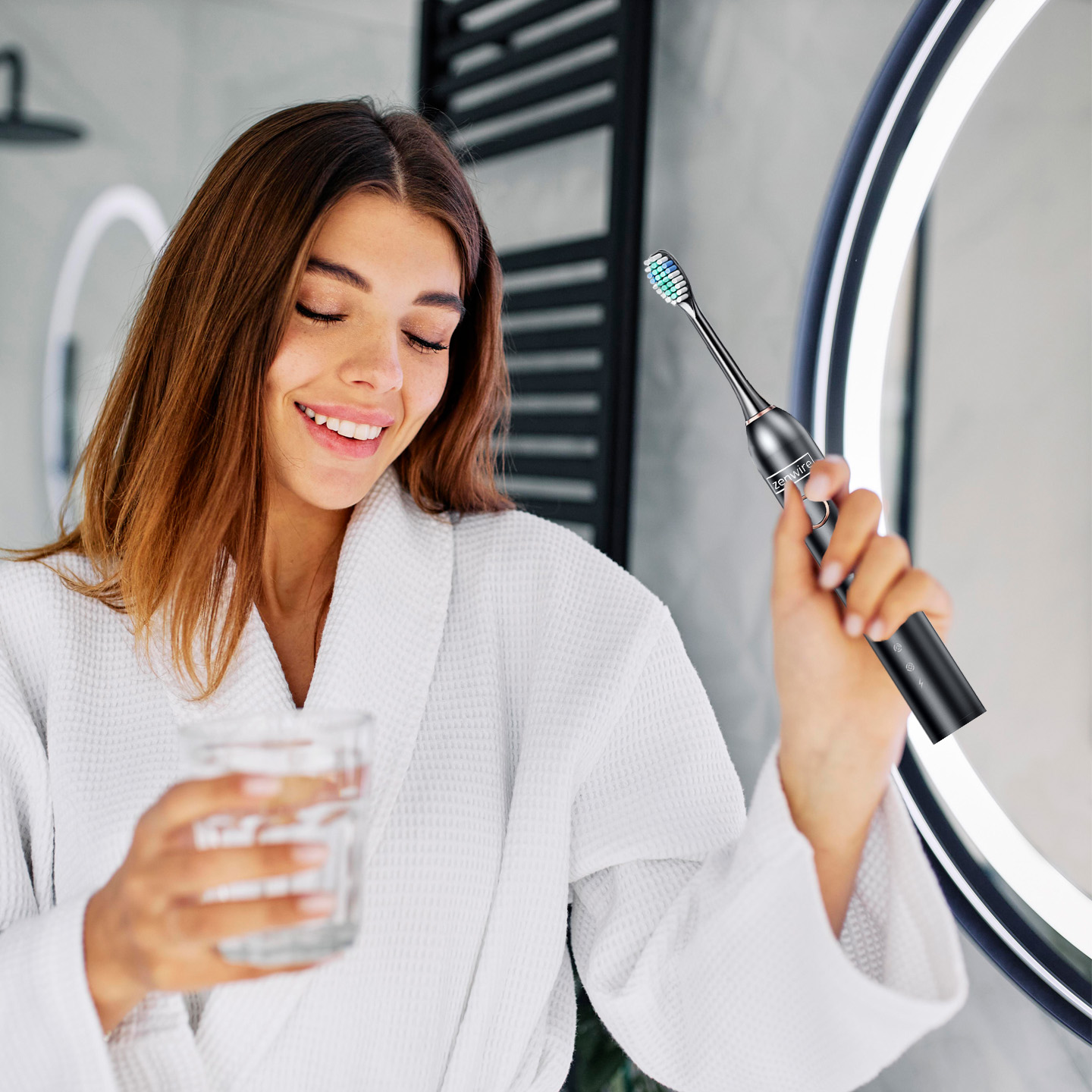 Kobieta w szlafroku uśmiecha się, trzymając szklankę i szczoteczkę do zębów w łazience z lustrem.