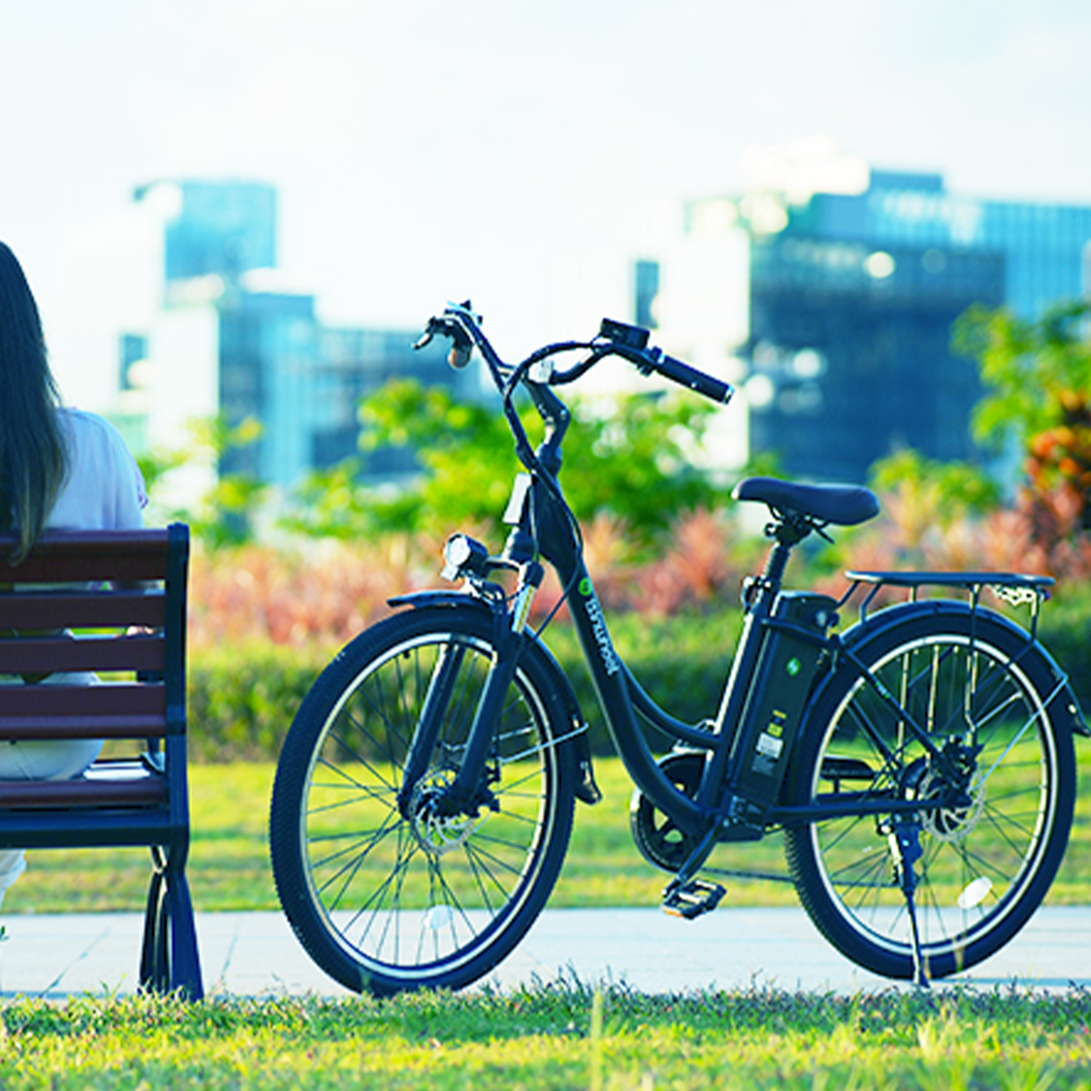 Czarny rower elektryczny zaparkowany obok ławki, z siedzącą osobą i budynkami w tle.