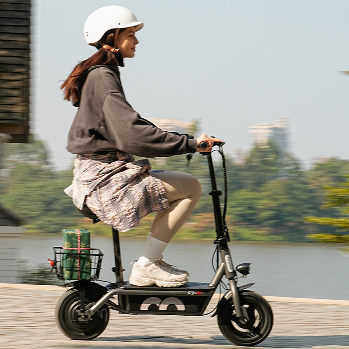 Kobieta na e-skuterze, w kasku, jadąca w pobliżu jeziora z prezentami w koszyku.