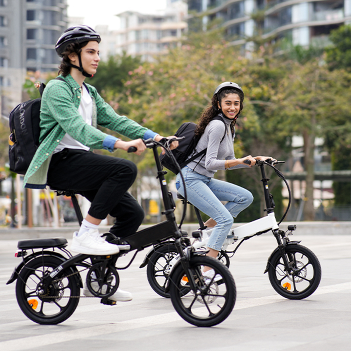 Dwie osoby jadące na składanych rowerach elektrycznych na ulicy miejskiej, uśmiechnięte, w kaskach.