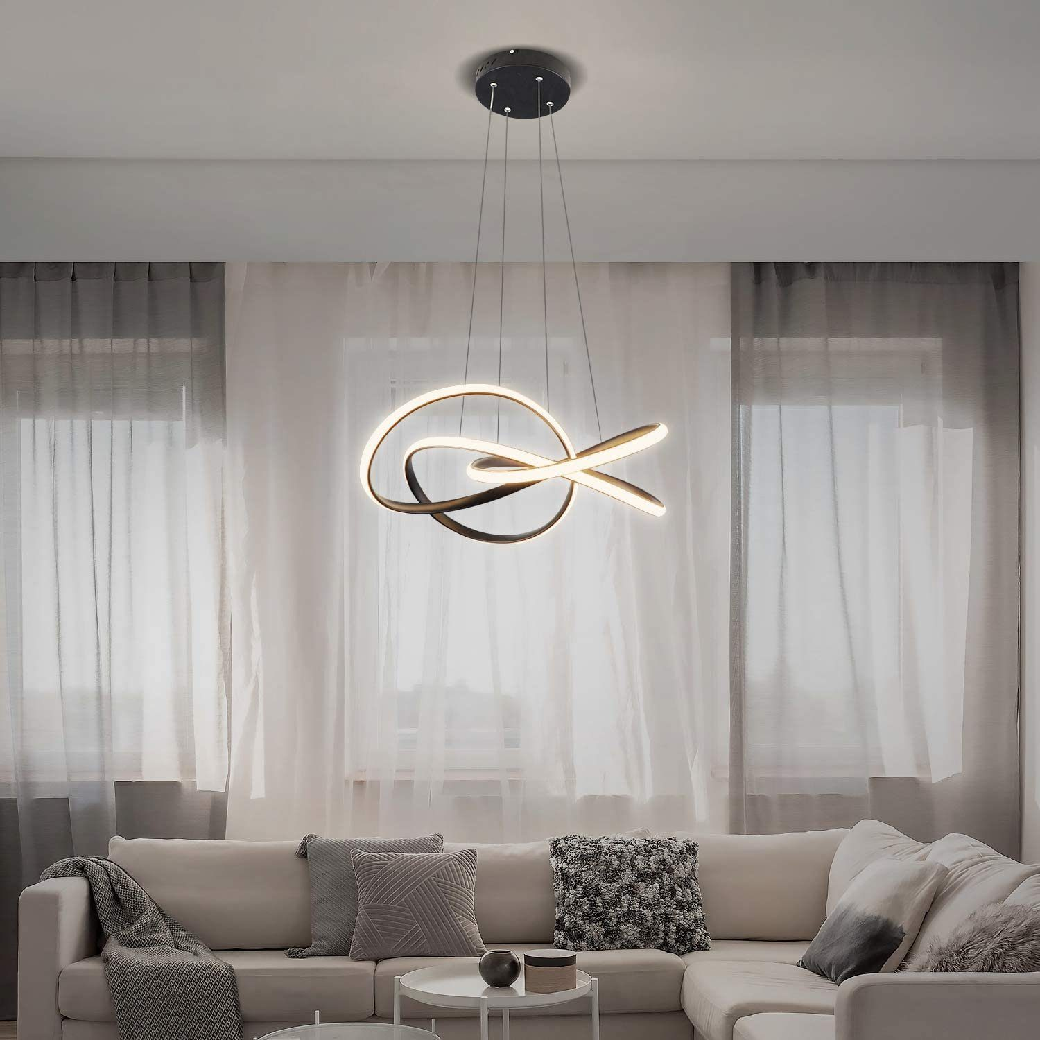 Moderne, verschlungene, goldfarbene Leuchte, die in einem Wohnzimmer hängt. Beige Sofa, transparente Vorhänge, graue Kissen.