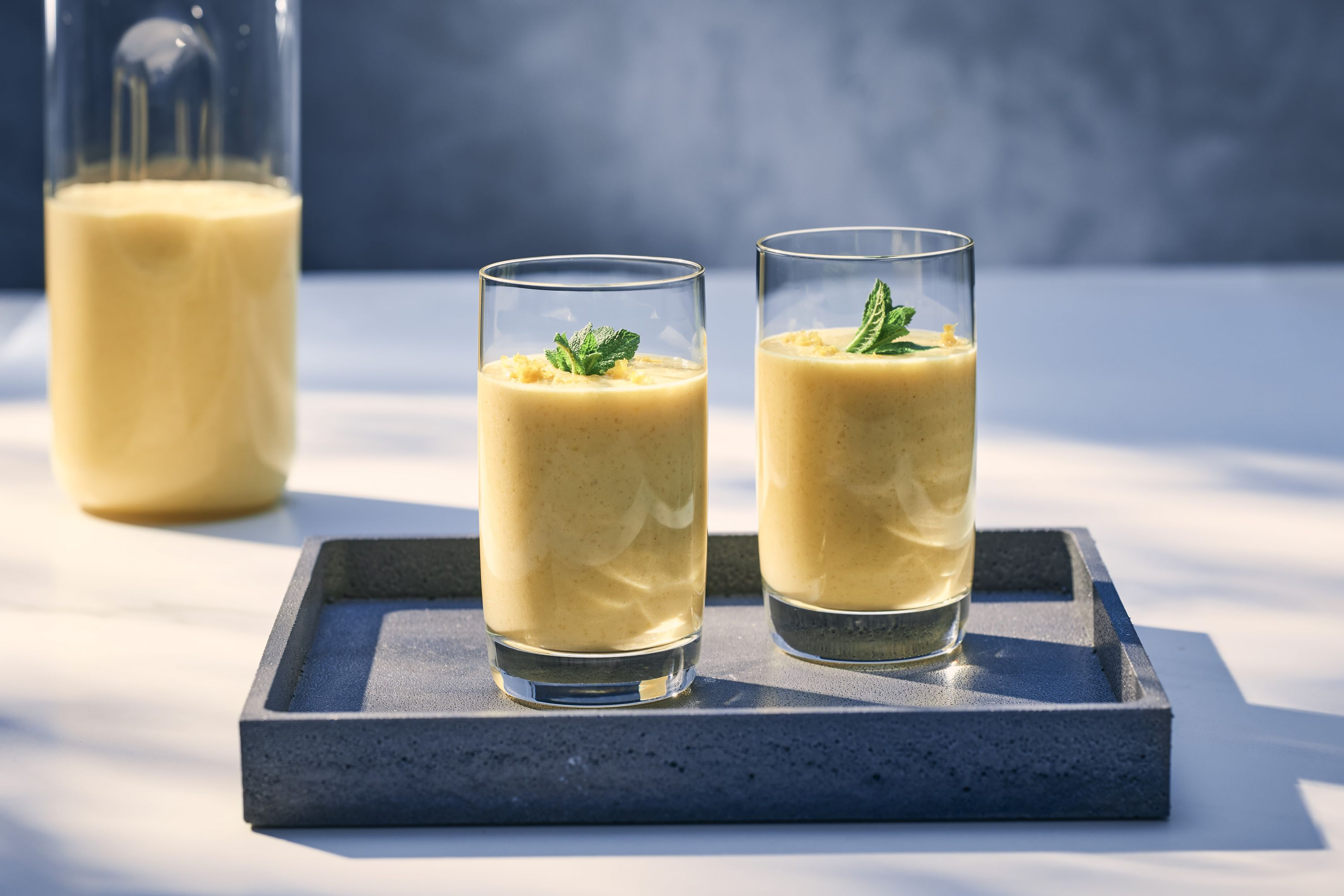 Zwei Gläser gelber Smoothie auf einem Tablett mit Minzdekor. Krug im Hintergrund.