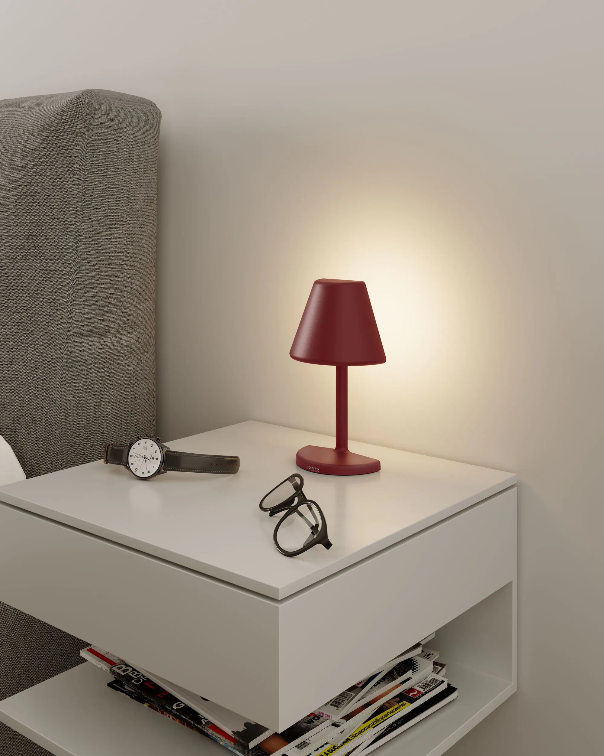 Una piccola lampada da comodino rossa su un comodino bianco, con occhiali e orologio.