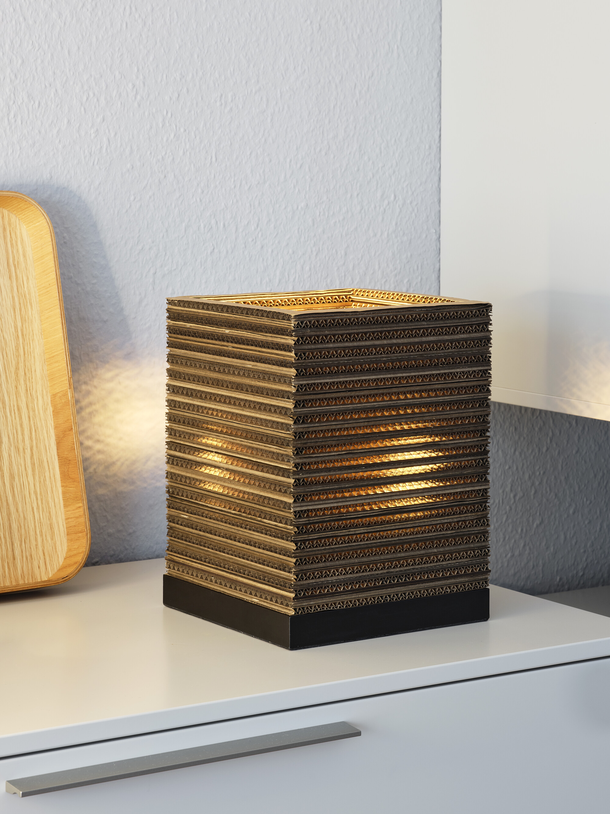 Eine würfelförmige Lampe aus brauner Wellpappe, mit schwarzem Sockel und Licht innen.
