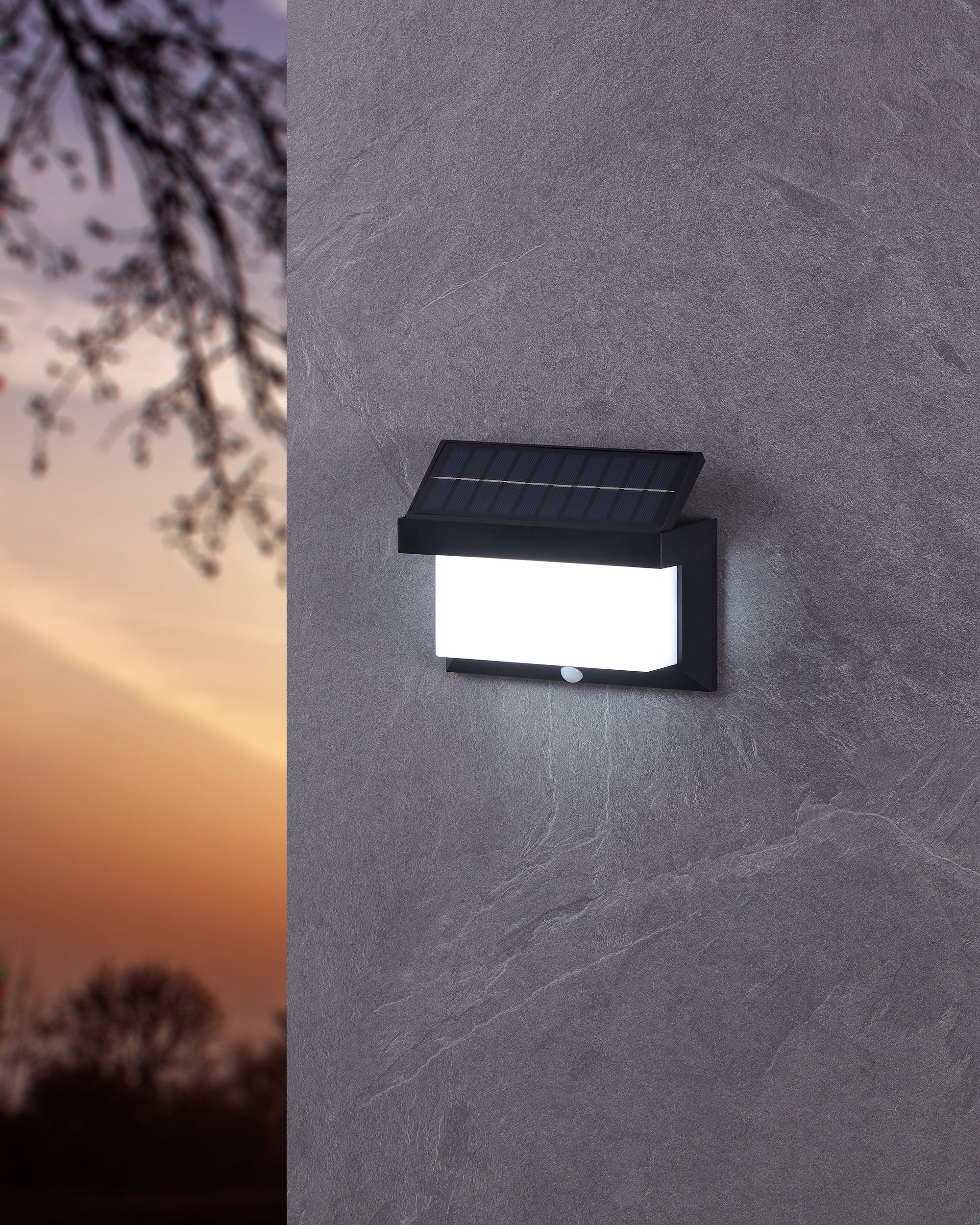 Lámpara de pared exterior con energía solar en una pared gris texturizada. Un atardecer en el fondo.