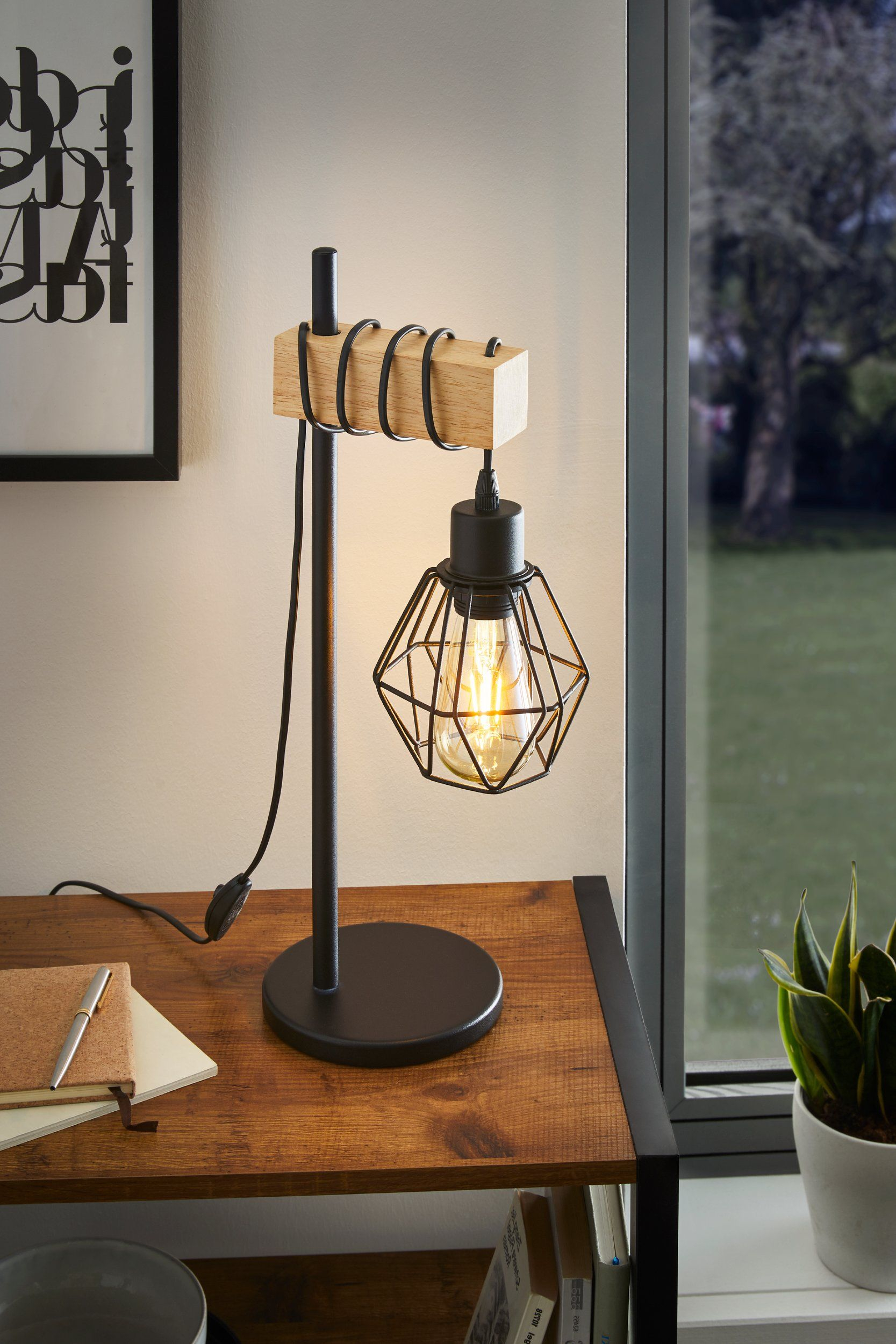 Lampada da tavolo su scrivania in legno, base in metallo nero, accento in legno. Finestra dietro.
