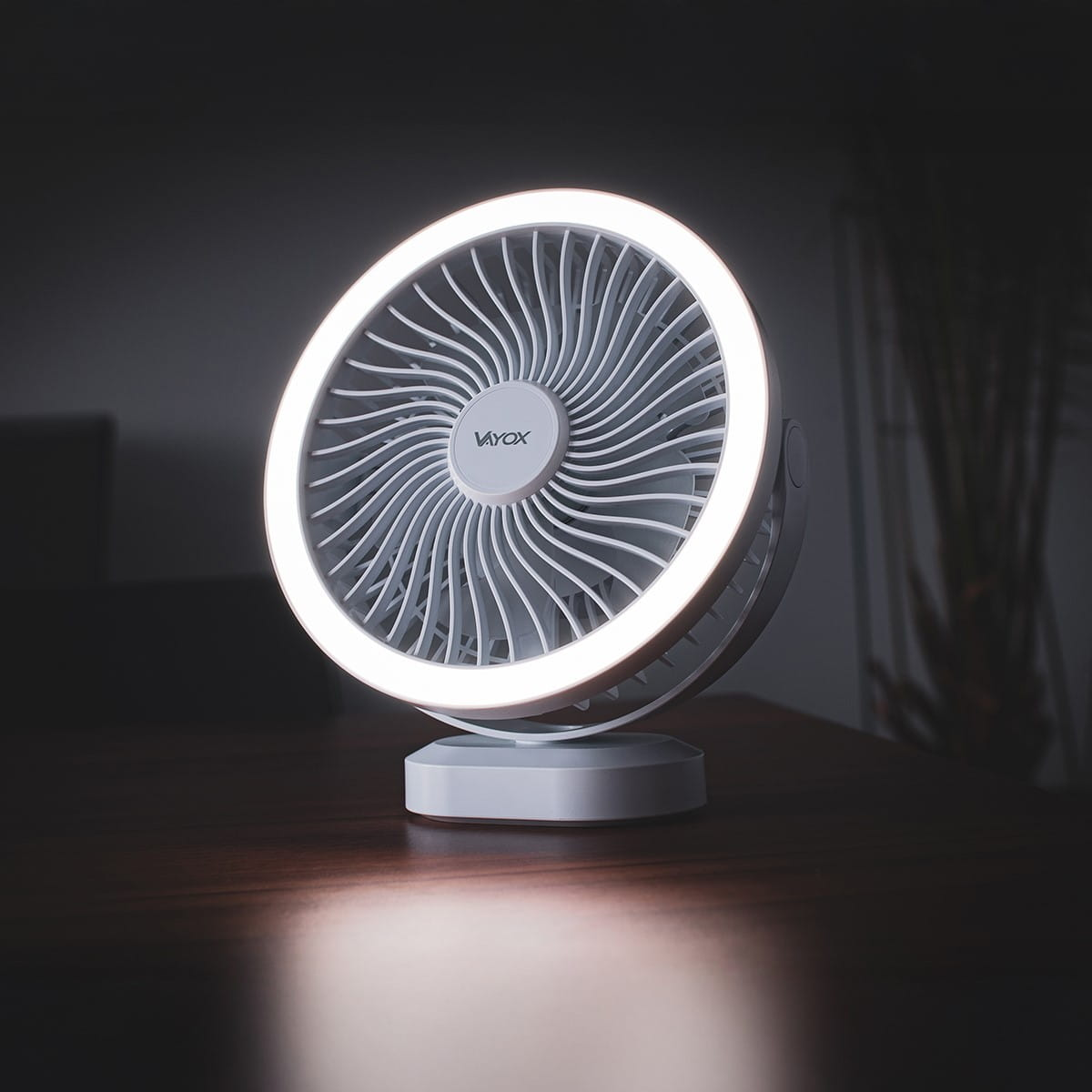 Biały wentylator z pierścieniem LED na ciemnym drewnianym stole. Widoczne logo VAYOX.