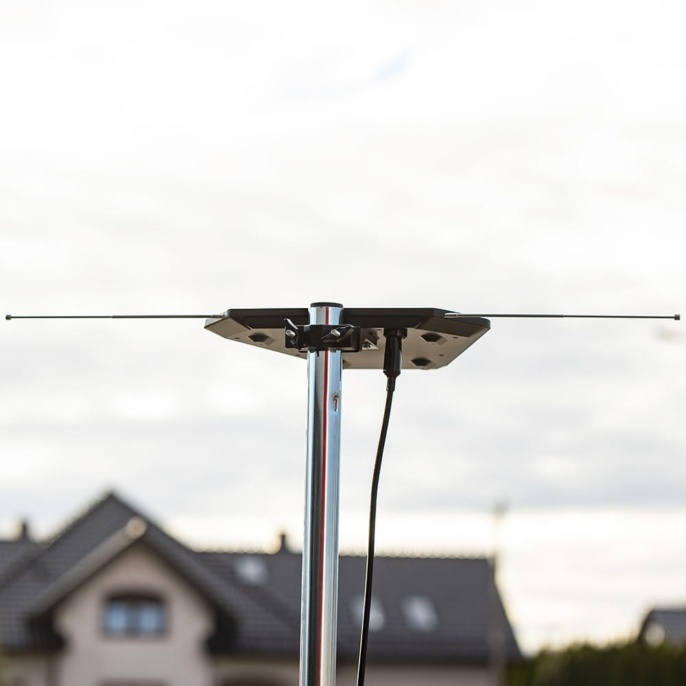 Czarna antena na metalowym słupie, z domem w tle i zachmurzonym niebem.