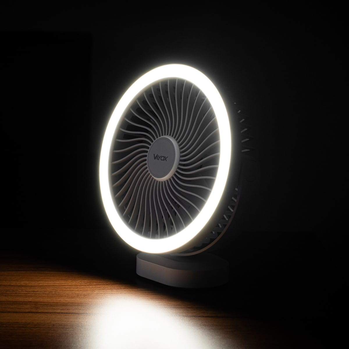 Szary wentylator z okrągłym światłem LED. Wentylator stoi na stole, oświetlony światłem.