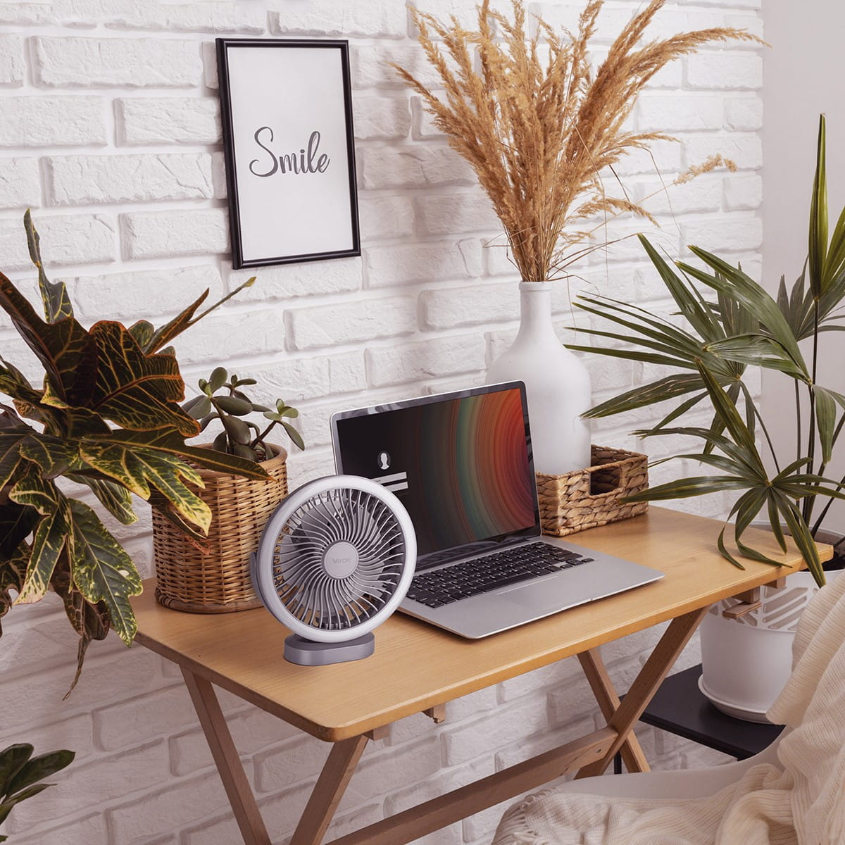 Biurko z laptopem, wentylatorem, roślinami i oprawionym znakiem 'Smile' na tle białej ceglanej ściany.