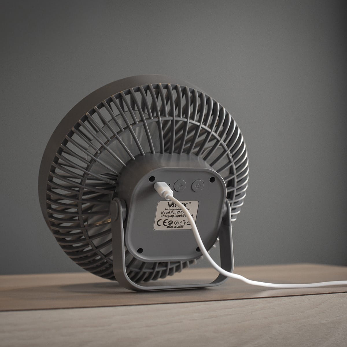 Szary wentylator biurkowy z boku, na drewnianym stole, z białym kablem.