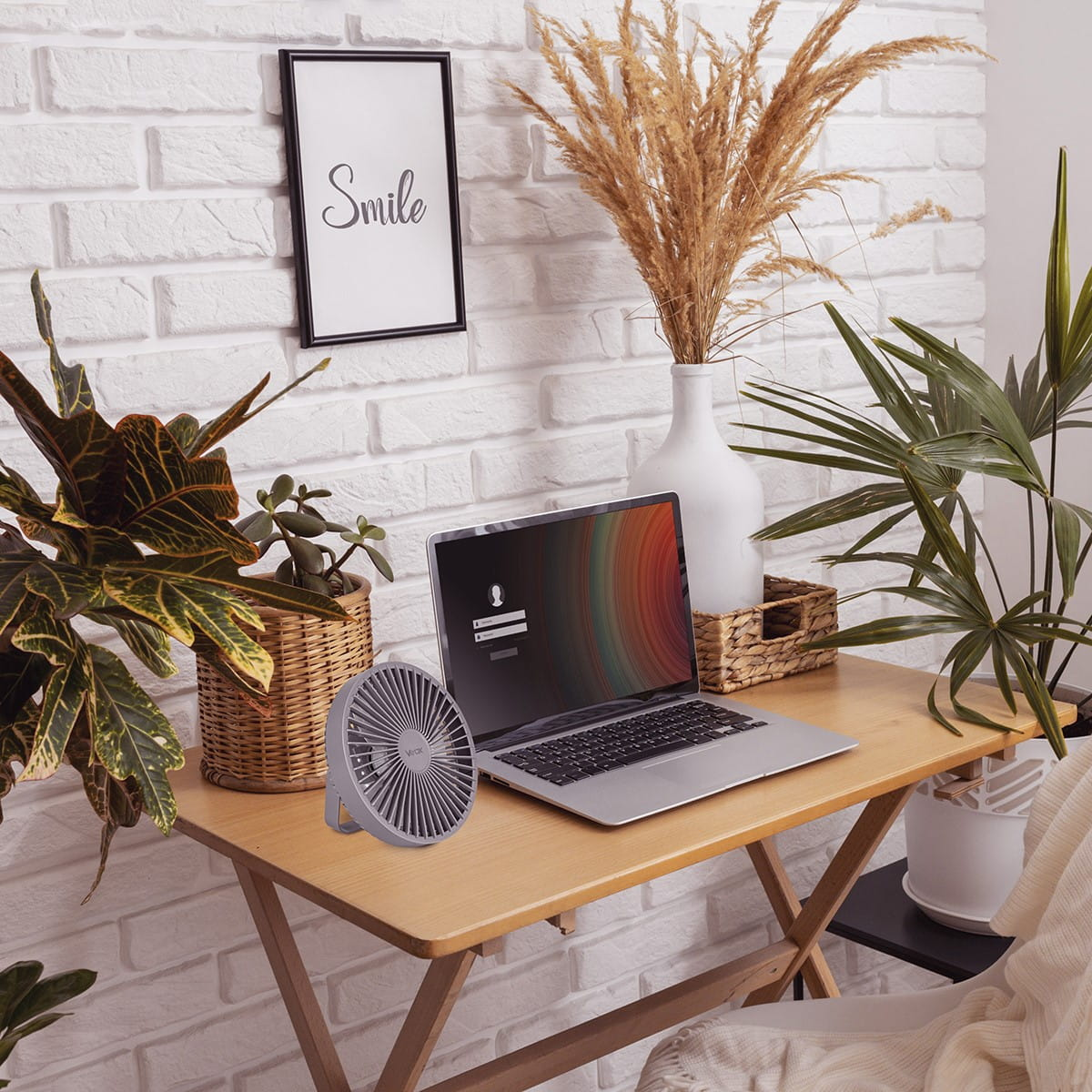 Konfiguracja biurka z laptopem, wentylatorem, roślinami i oprawioną grafiką na tle ceglanej ściany.