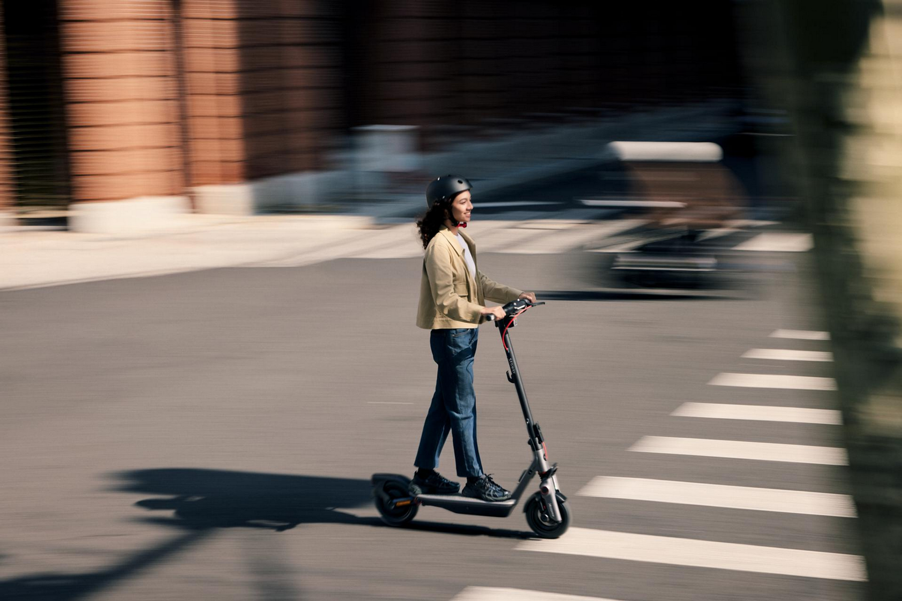 Eine Frau fährt mit einem Elektroroller auf einem Zebrastreifen und trägt einen Helm.
