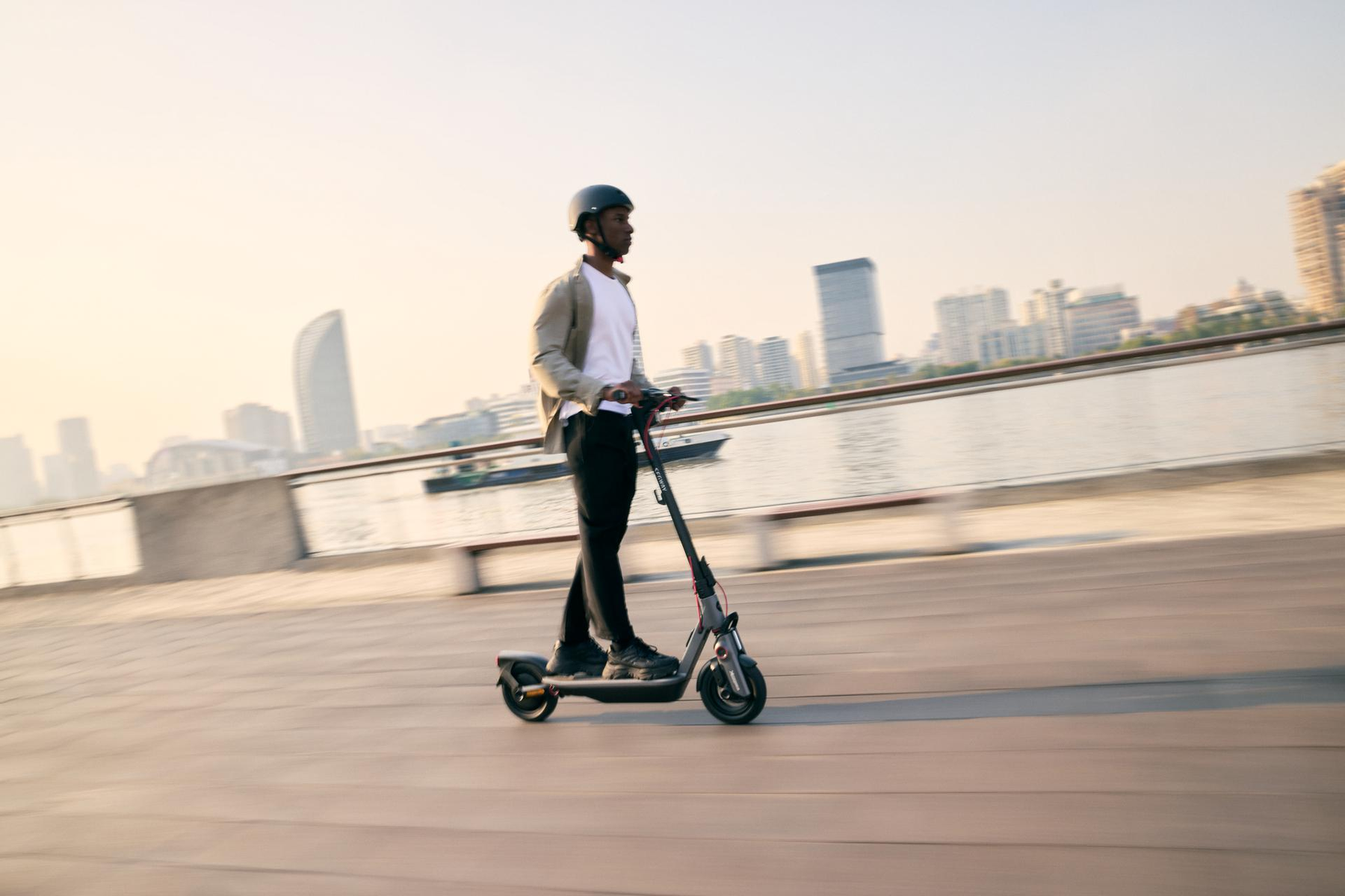 Un hombre con casco monta un scooter en un día soleado, cerca de edificios y una carretera.