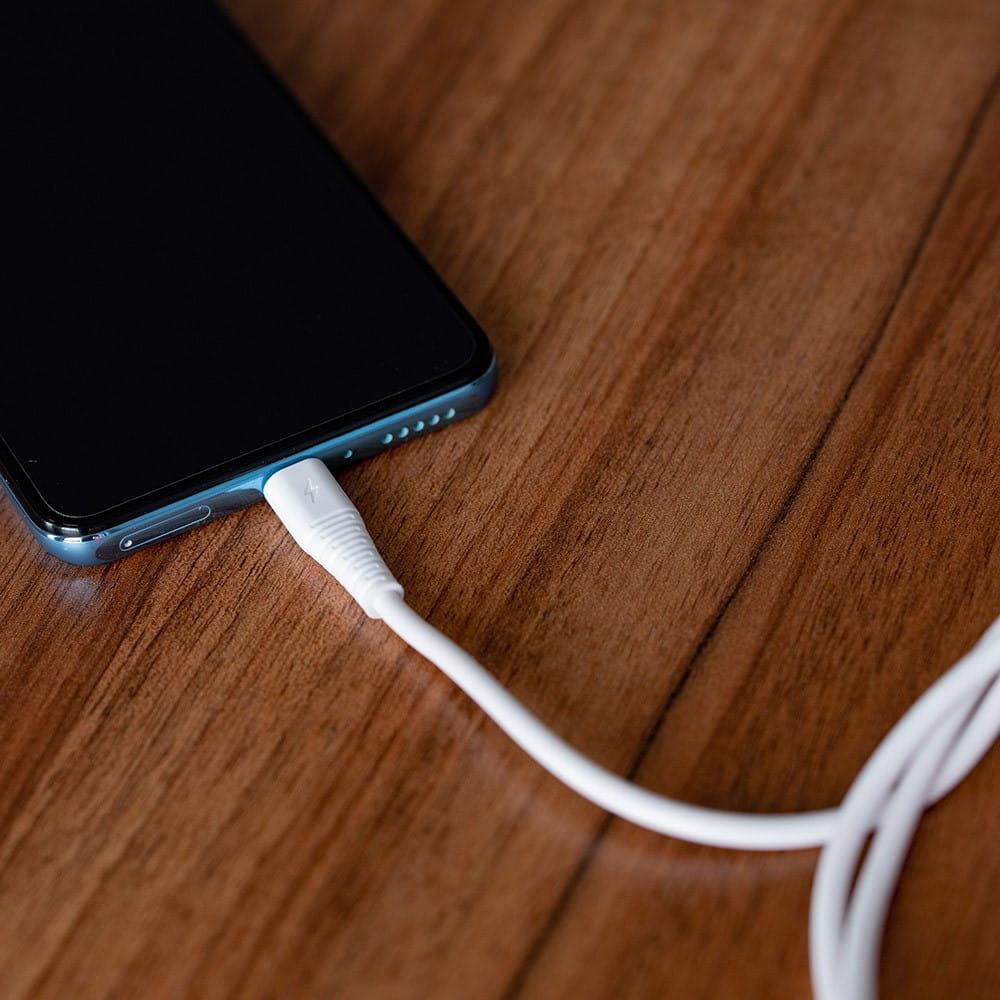 Smartfon z podłączonym białym kablem ładującym. Telefon leży na drewnie.