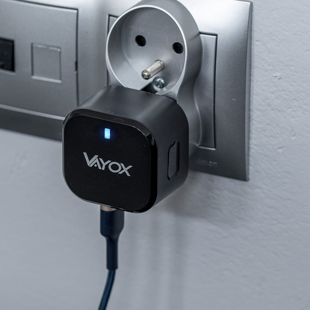 Czarna ładowarka Vayox podłączona do ściany. Świeci się kontrolka ładowania. Srebrne i białe gniazdo ścienne.