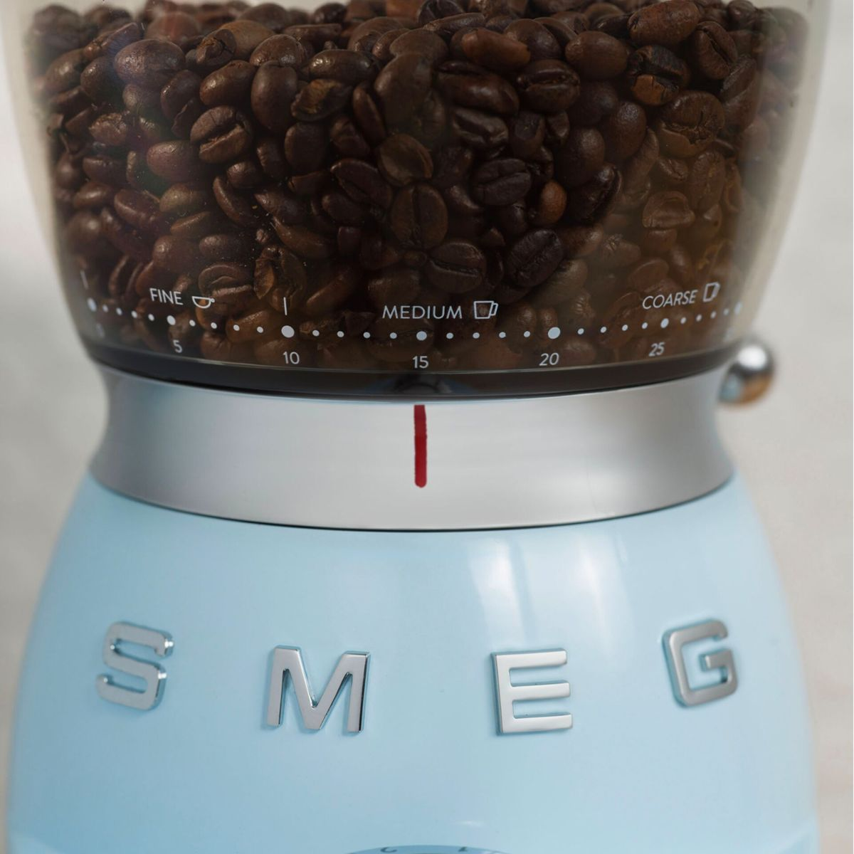 Gros plan sur un moulin à café Smeg bleu clair avec des grains de café dans la trémie. Le moulin est sur une surface.