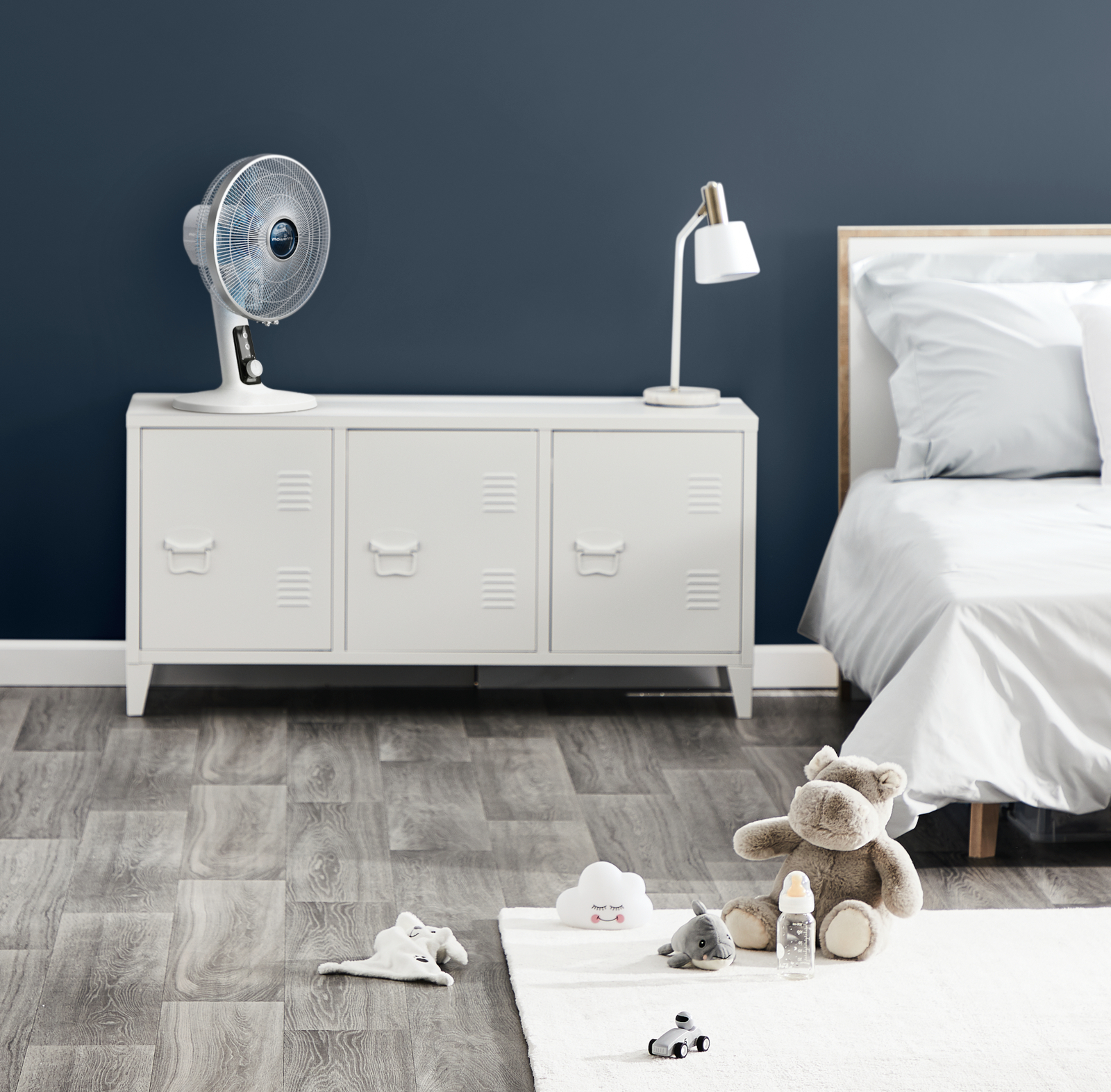 Scena sypialni z białą szafką, wentylatorem, lampą, zabawkami i łóżkiem. Ściana jest ciemnoniebieska.