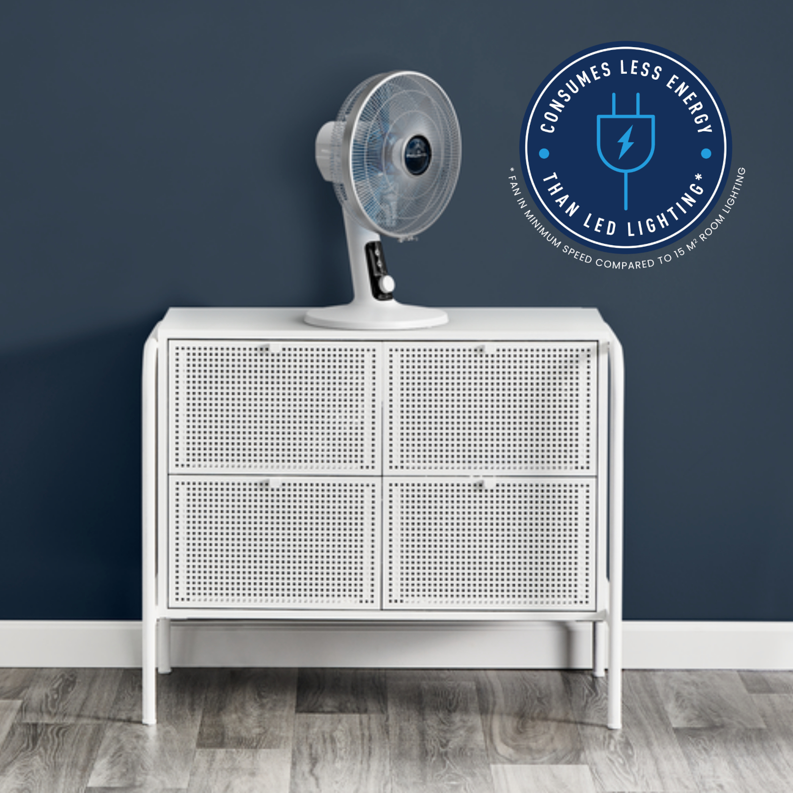 Biały wentylator na białej szafce na tle niebieskiej ściany. Tekst: Zużywa mniej energii niż oświetlenie LED.