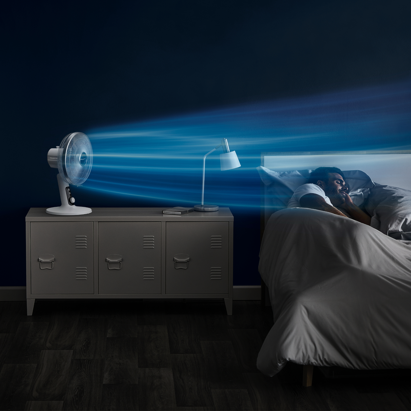 Mężczyzna śpiący w łóżku z wentylatorem dmuchającym w jego kierunku. Pokój ma ciemne ściany i stolik nocny.