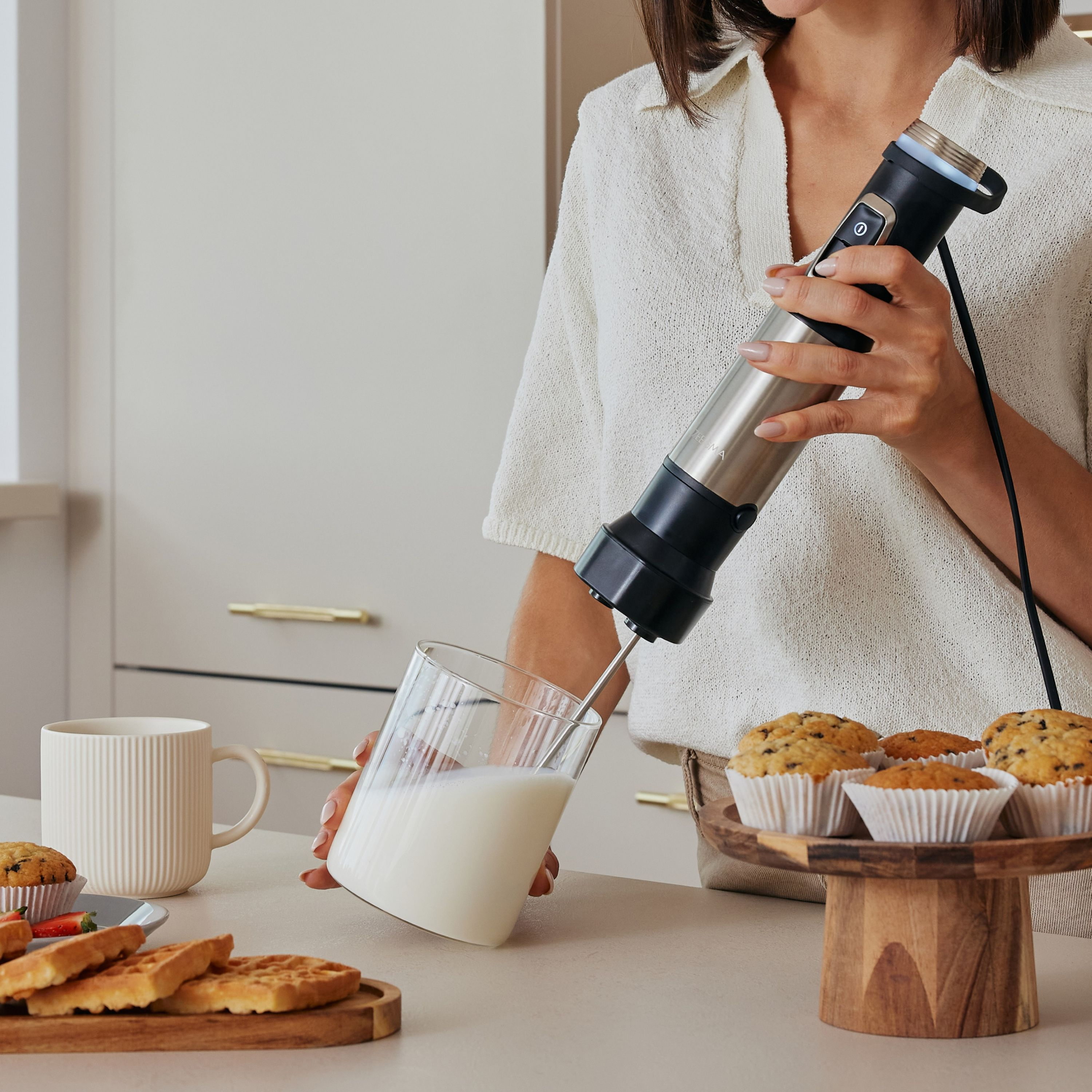 Eine Frau benutzt einen Milchaufschäumer. Milch wird in ein Glas gegossen, neben Waffeln, Muffins und einer Tasse.