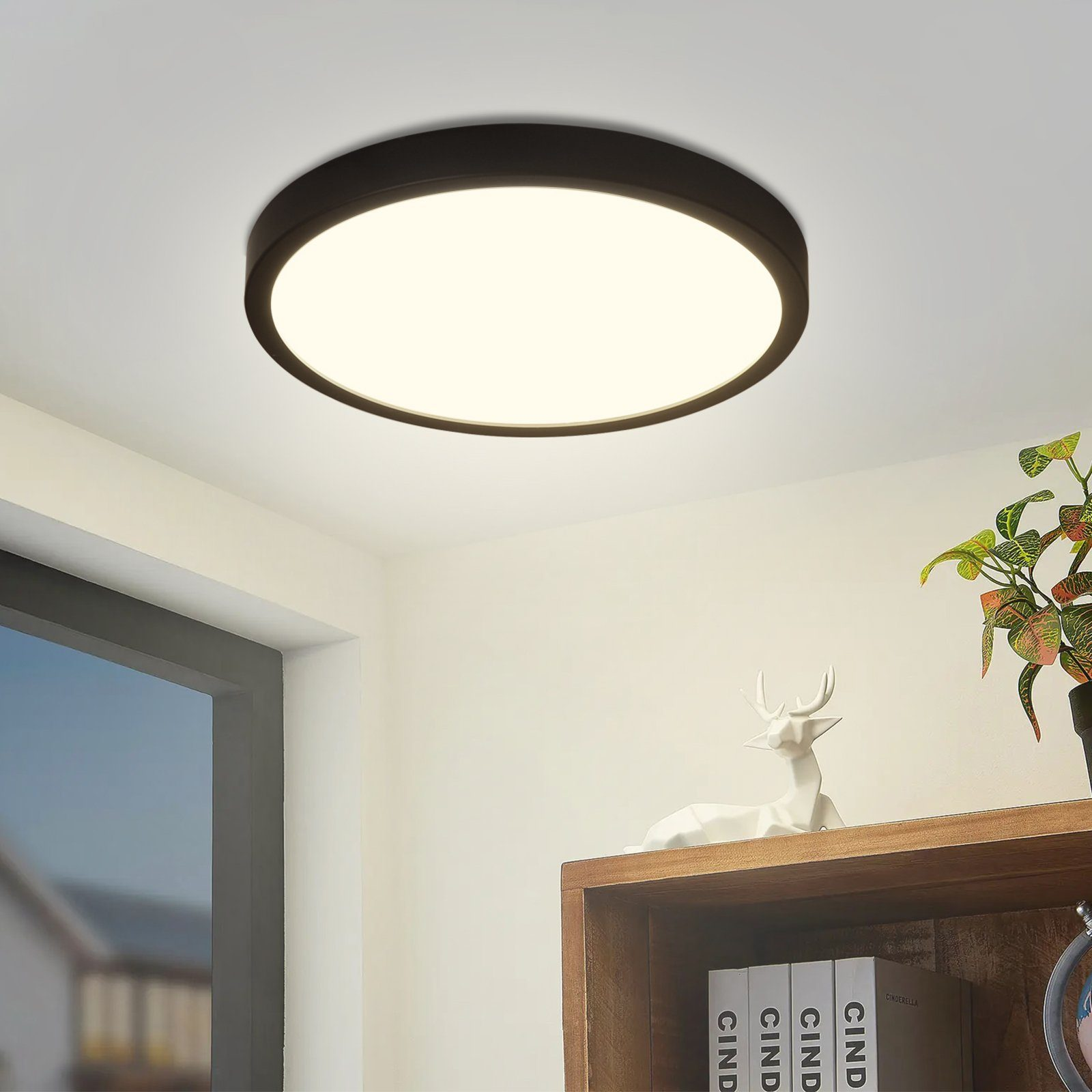 Runde LED-Deckenleuchte in einem Raum. Schwarzer Rahmen, weißes Leuchtpanel. Dekorative Elemente im Hintergrund.