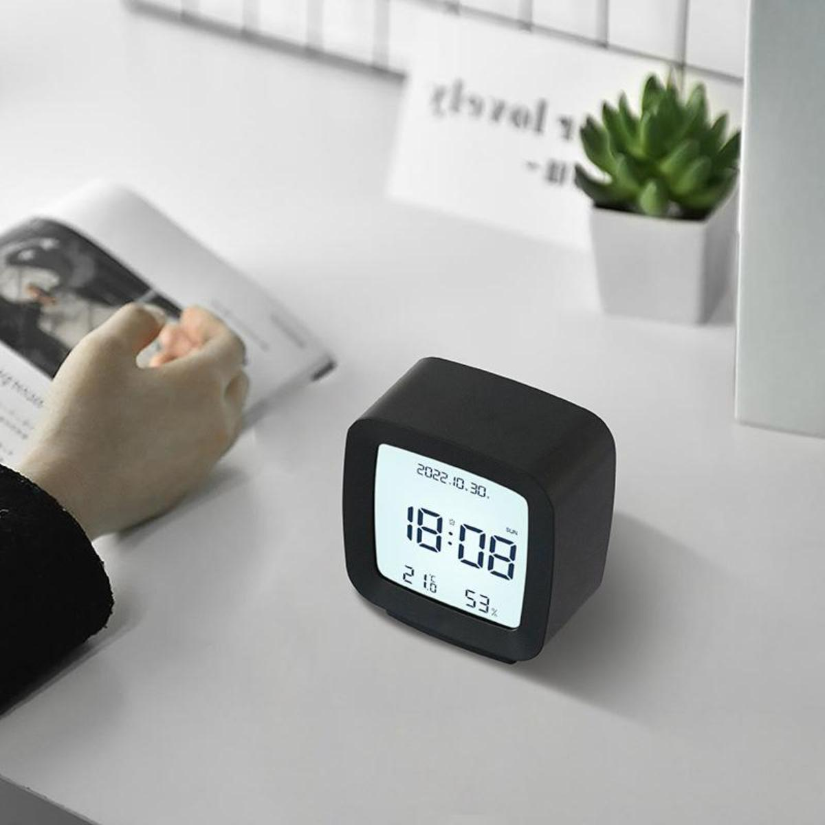 Schwarzer digitaler Wecker auf weißer Oberfläche. Zeigt Zeit, Datum, Temperatur und Luftfeuchtigkeit.