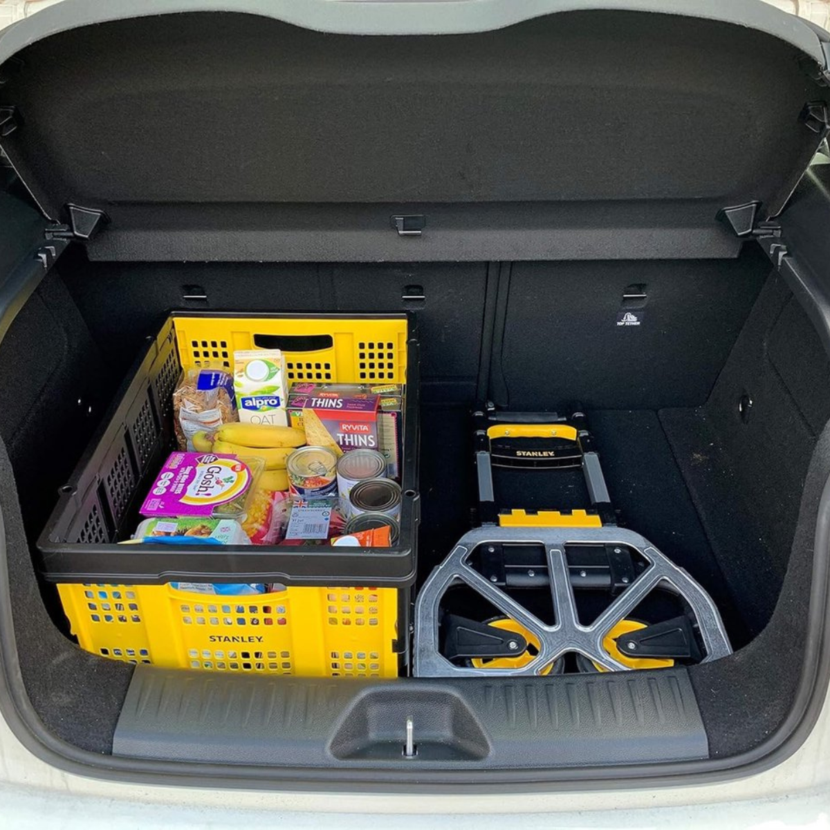 Car trunk with groceries in yellow and black crates, and a foldable hand truck.