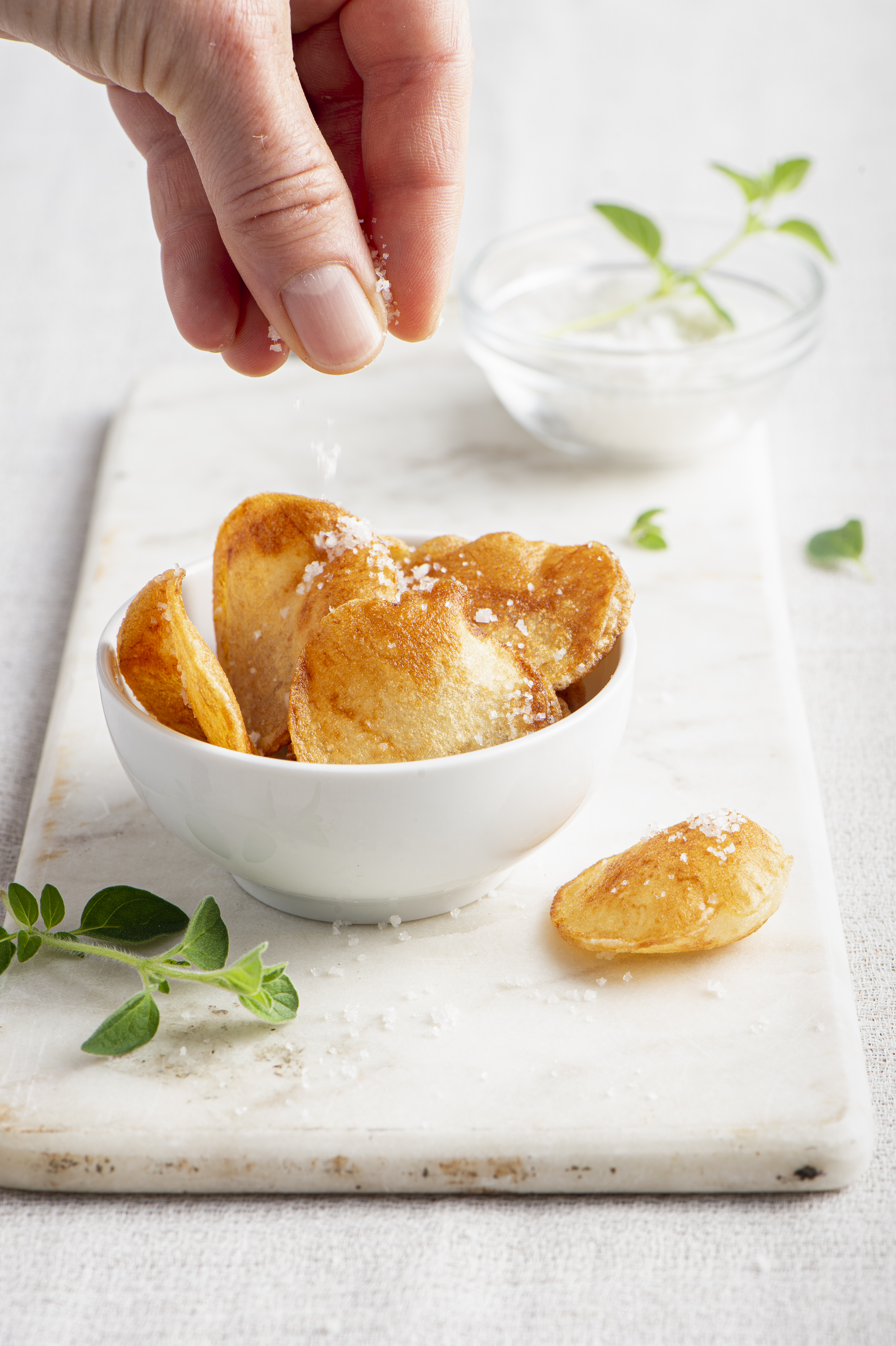 Hand sprinkling salt onto a bowl of potato chips, with a small bowl of salt and oregano.