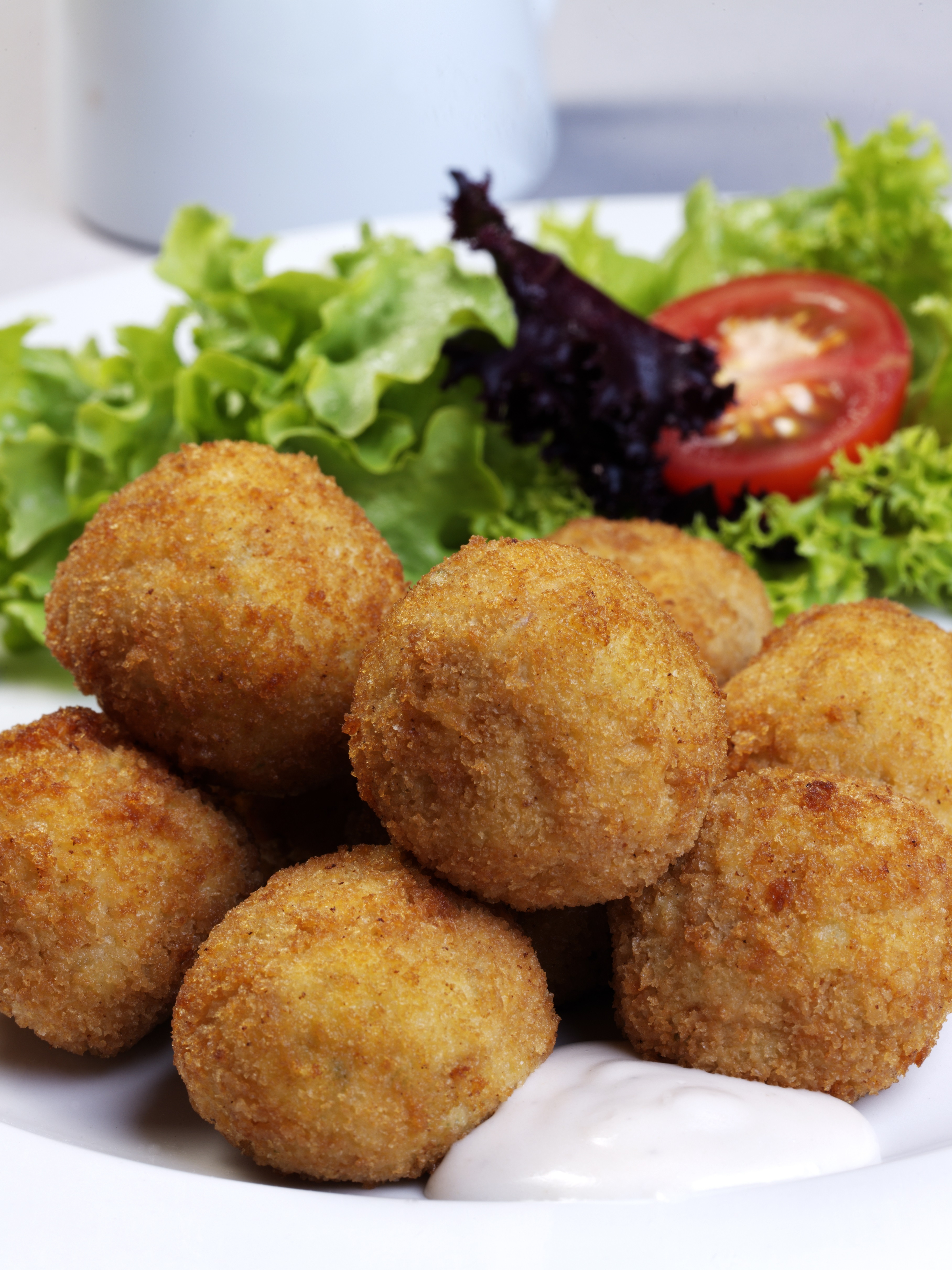 Several golden-brown fried croquettes on a white plate, served with greens and a tomato.