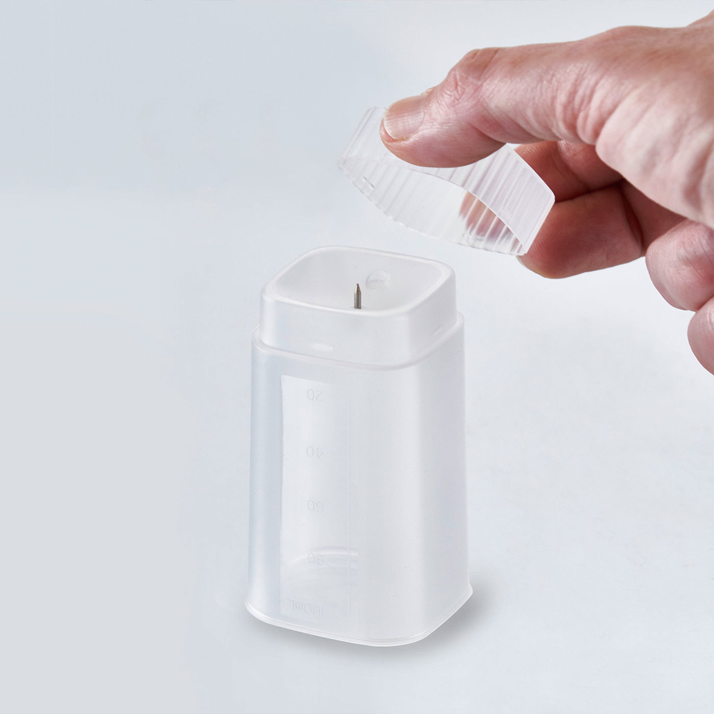 Dłoń trzyma przezroczysty plastikowy pojemnik z podziałką. Zdejmowana pokrywka.