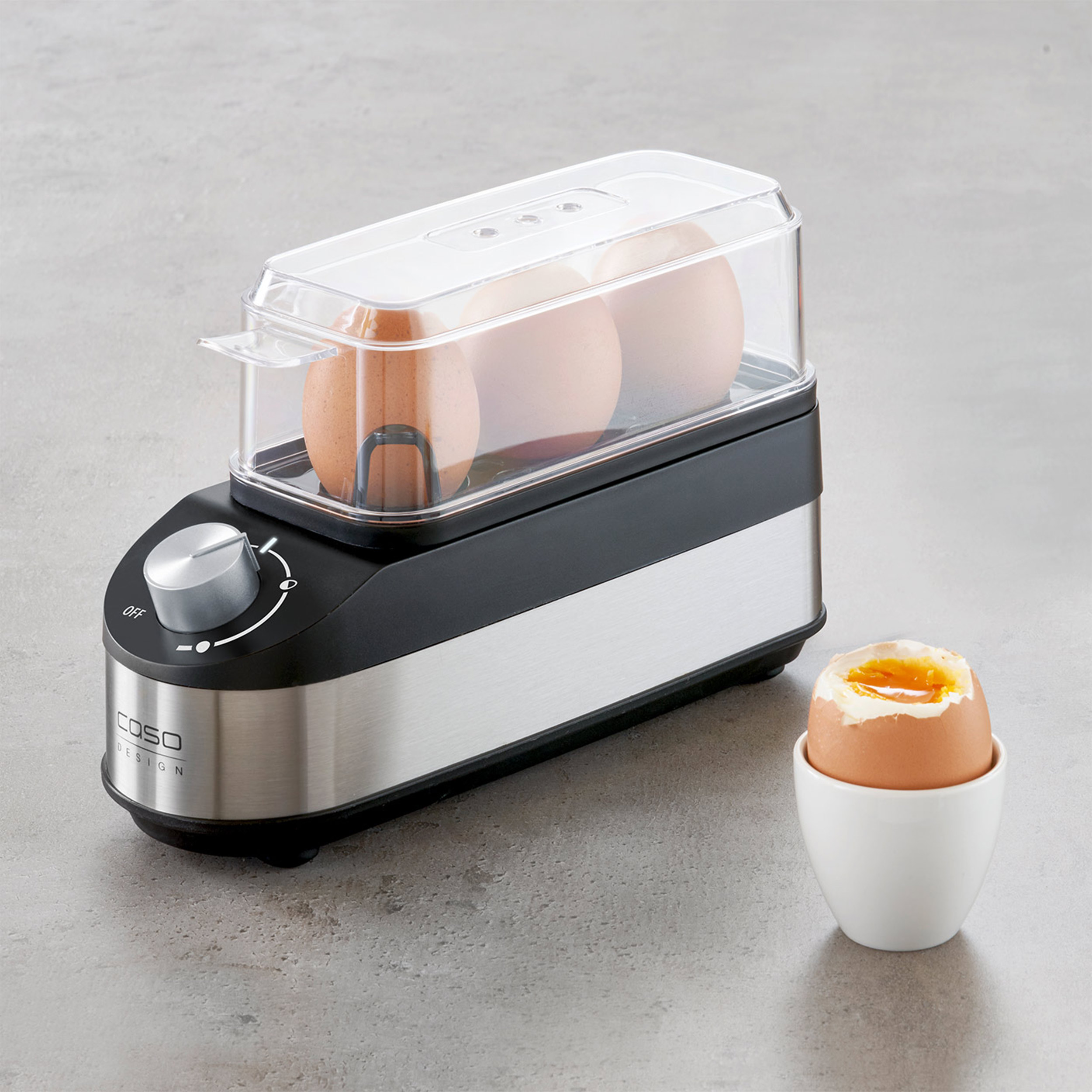 Gotowarka do jajek z trzema jajkami w środku. Jedno jajko siedzi w białym kubku, otwarte.