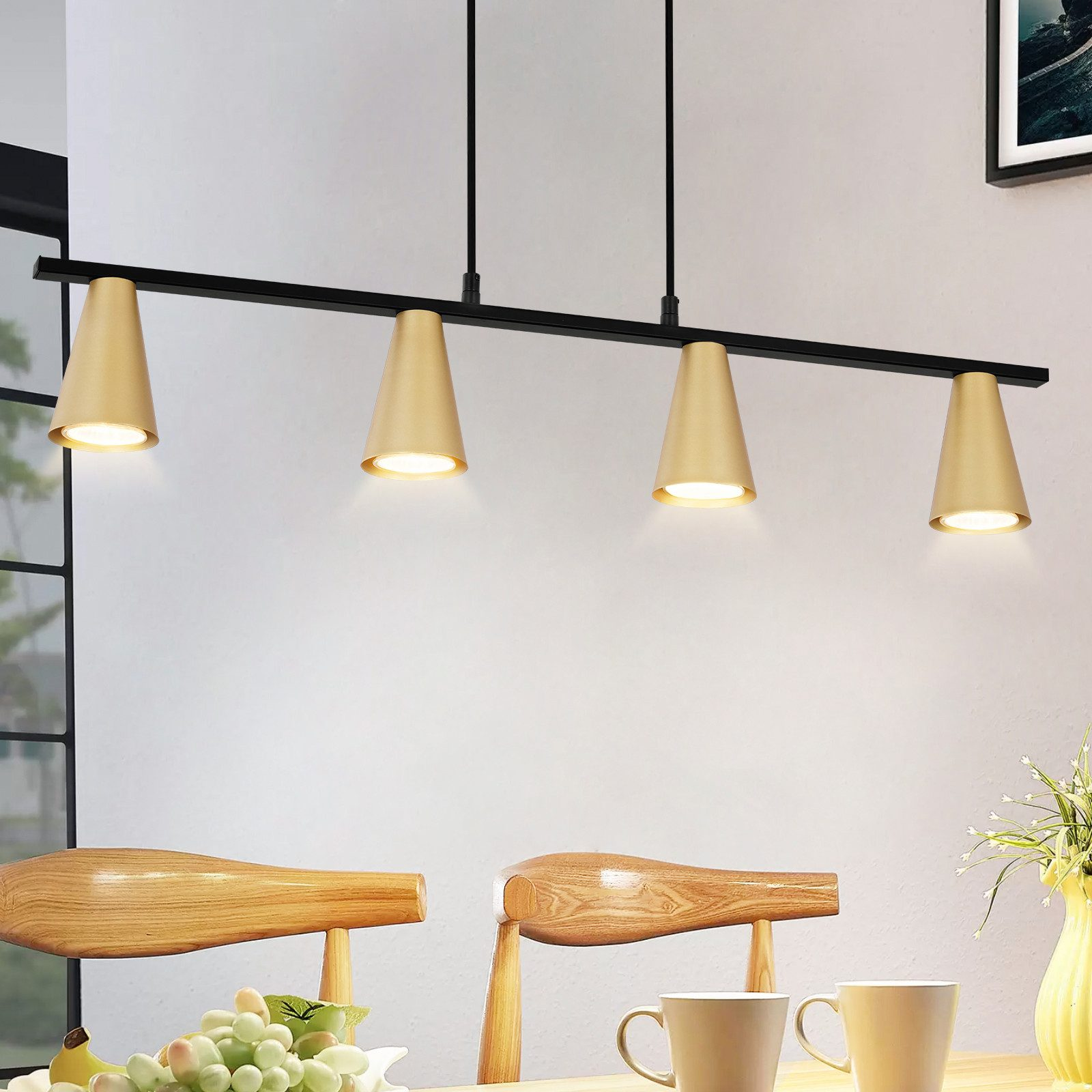 Lampada da soffitto con quattro lampade a forma di cono dorato. Sedie e tavolo in legno in primo piano. Sfondo parete bianca.