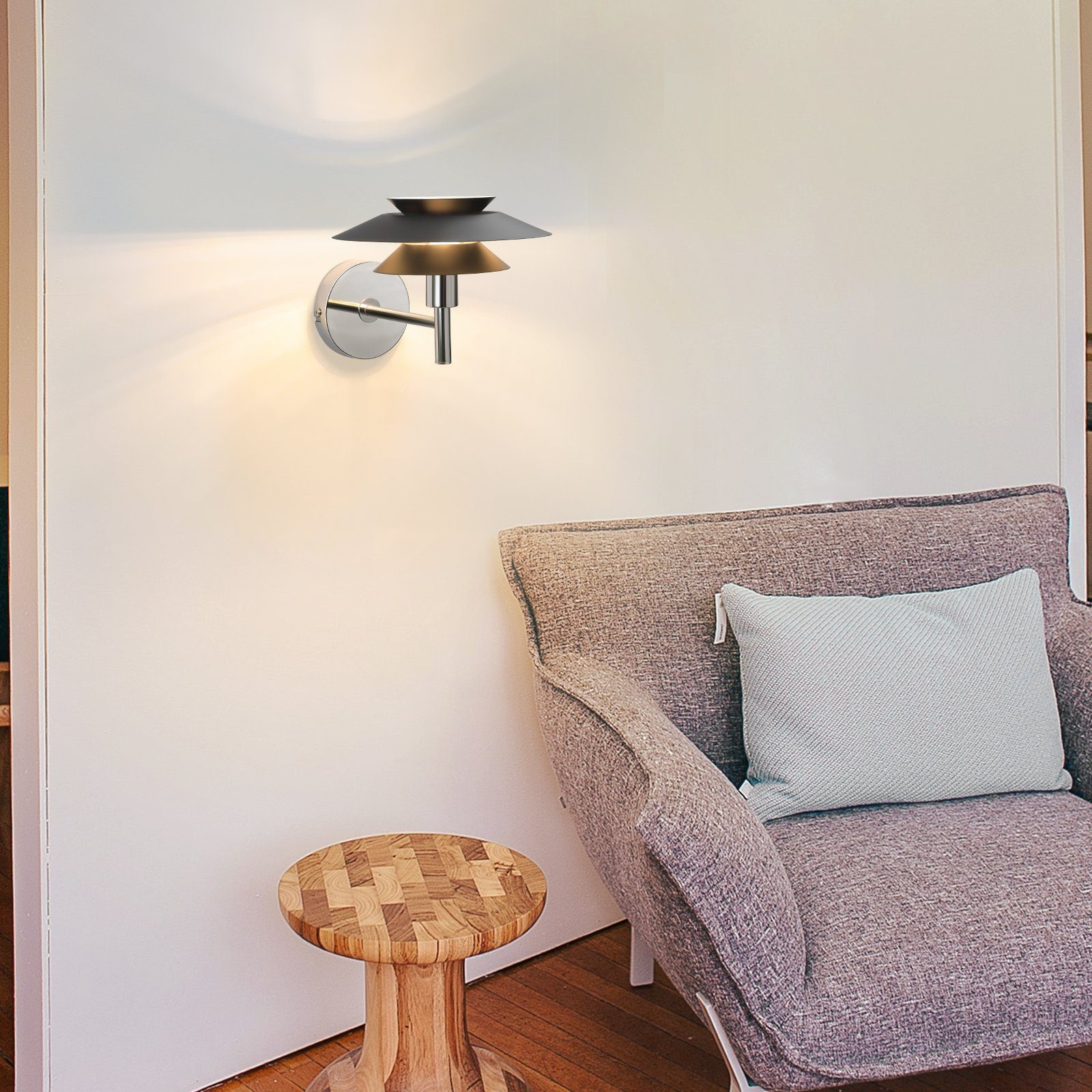 Lampada da parete con base argentata e paralume nero. È montata su un muro bianco.