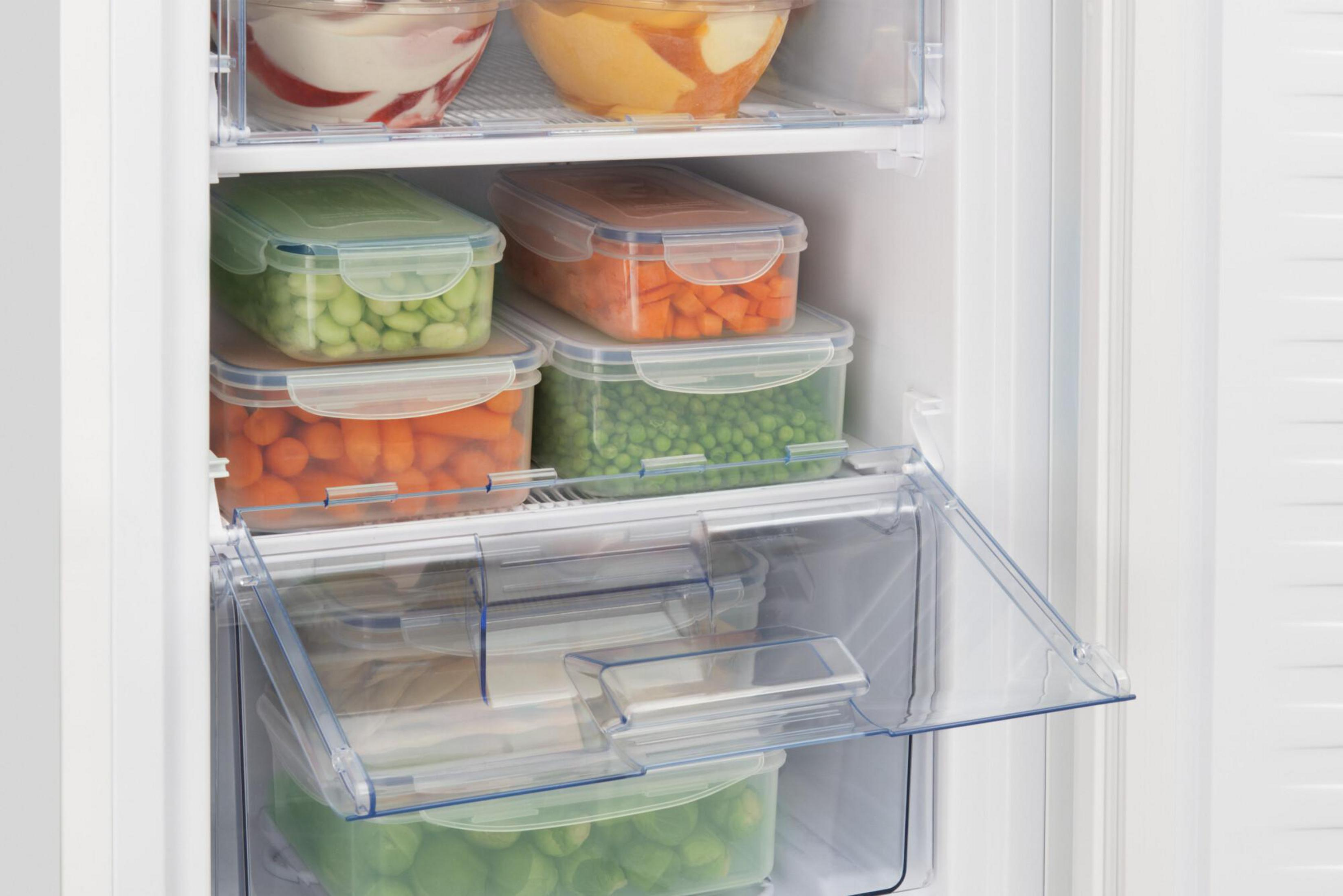 Zamrażarka z otwartymi drzwiami, pokazująca zamrożoną żywność w przezroczystych pojemnikach, w tym warzywa i gotowe dania.