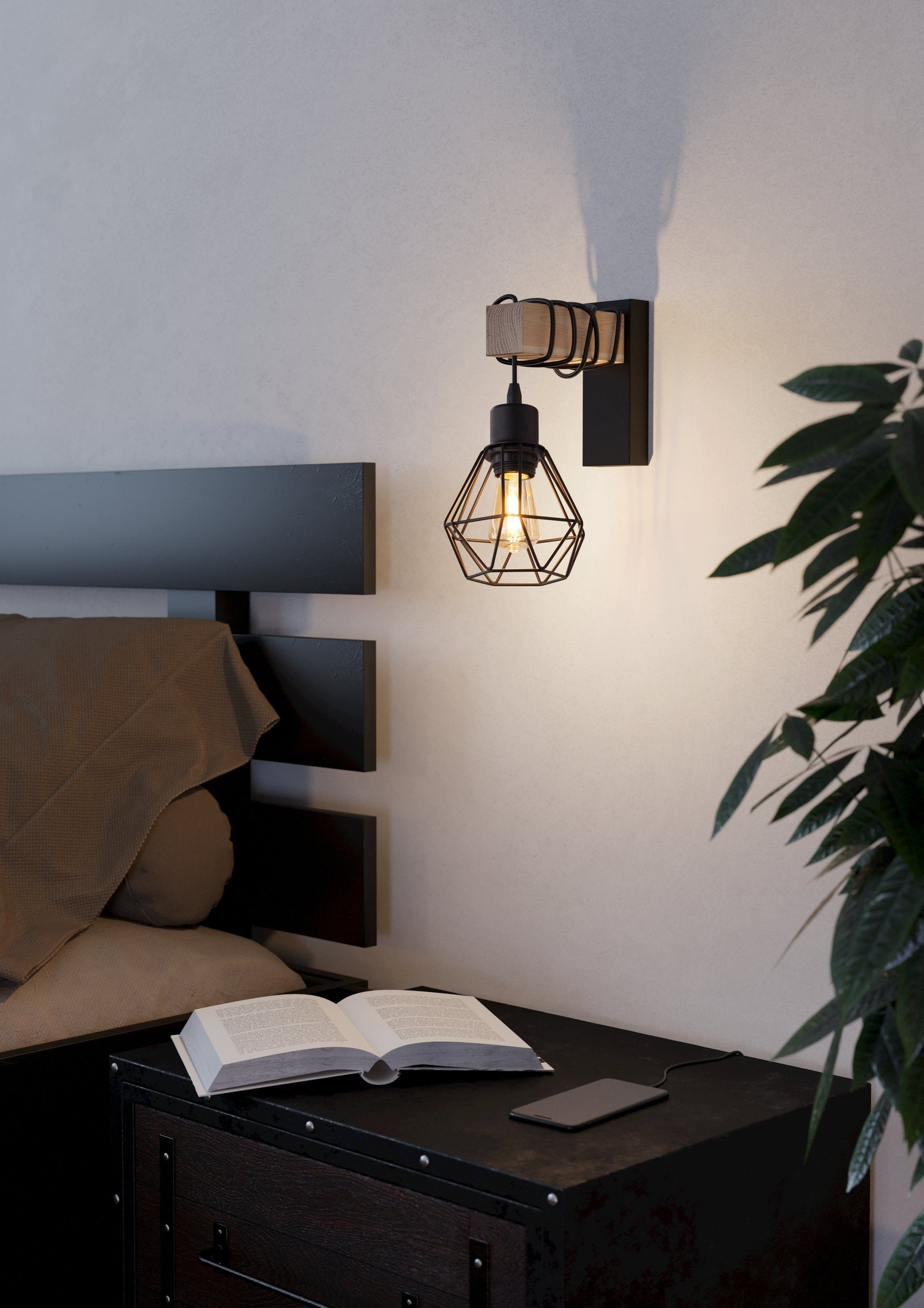 Una scena da comodino: lampada con lampadina accesa, libro, telefono e pianta, in una stanza.