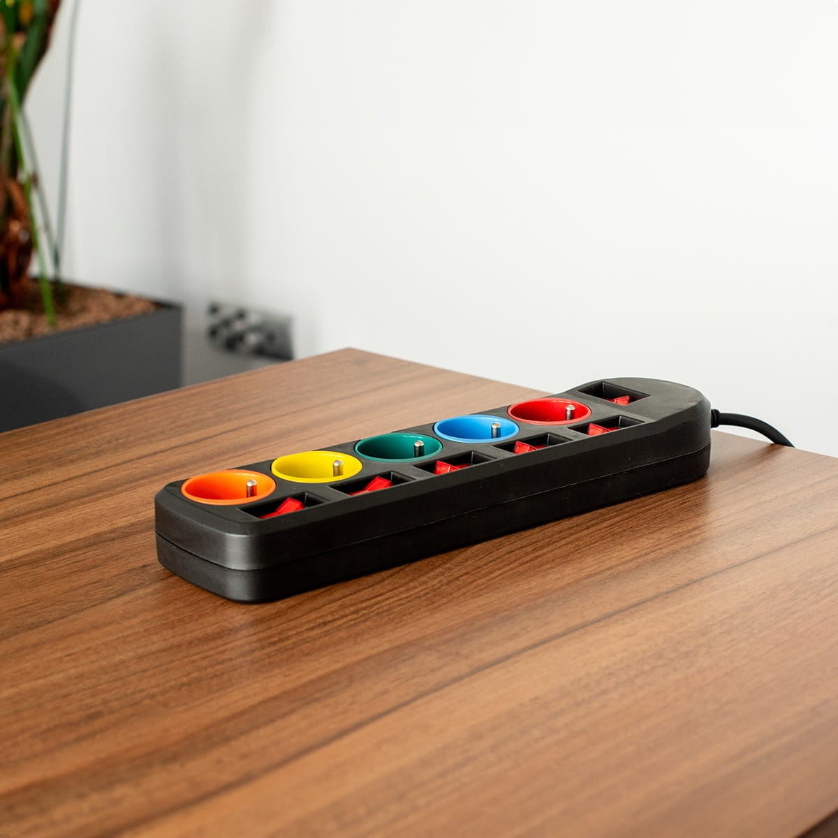 Czarna listwa zasilająca z sześcioma kolorowymi gniazdami na brązowym drewnianym stole.