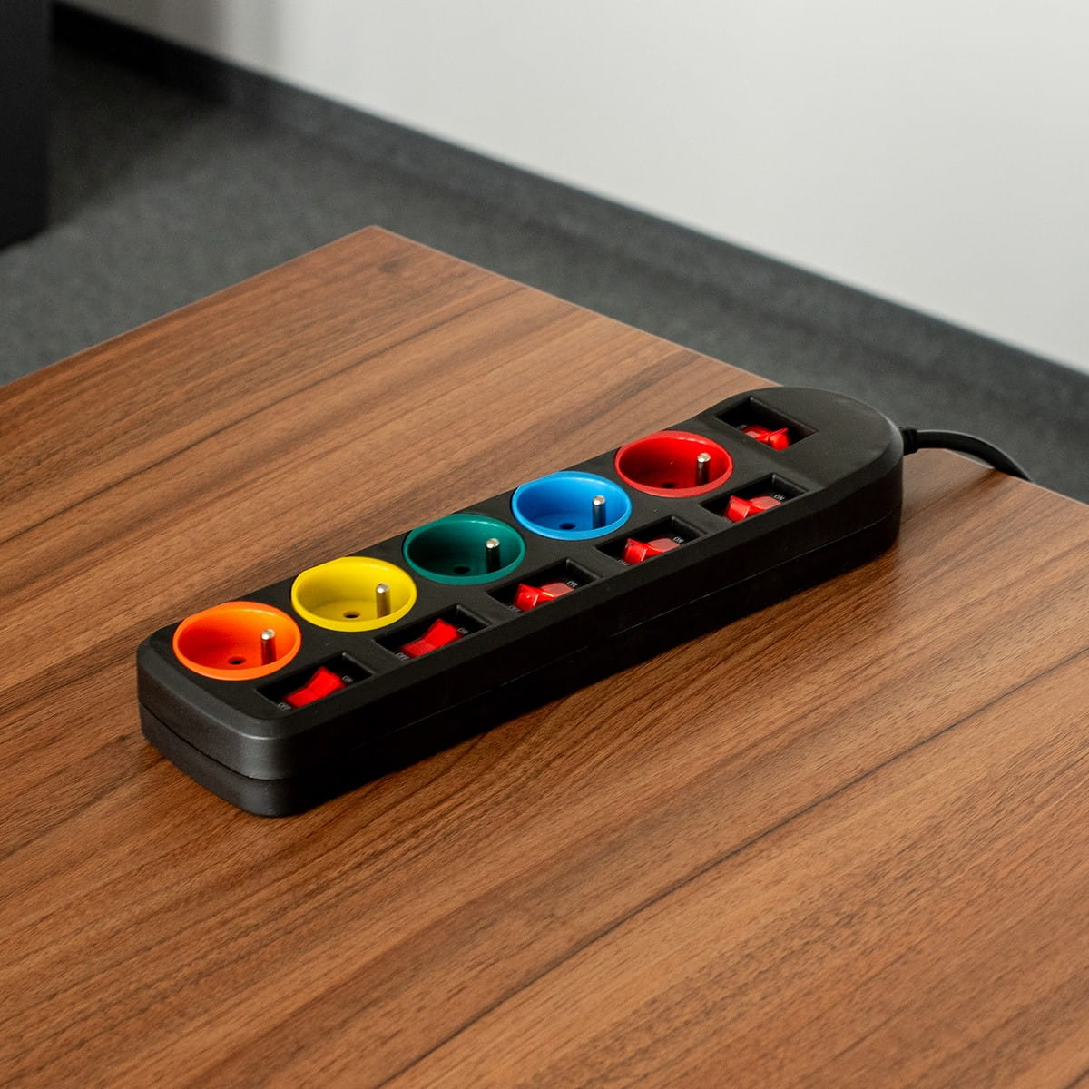Listwa zasilająca z pięcioma kolorowymi gniazdami i czerwonymi przełącznikami, na drewnianej powierzchni.