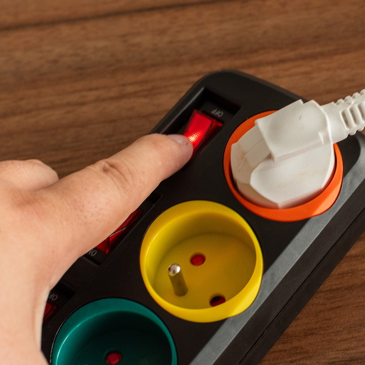 Ręka naciska czerwony przełącznik na listwie zasilającej z częściowo włożoną białą wtyczką.