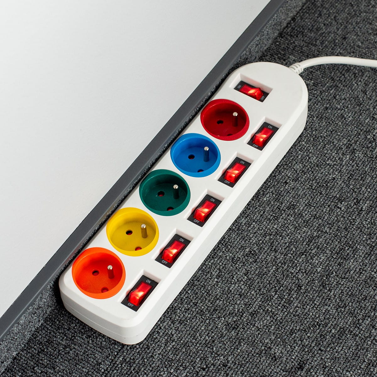 Biała listwa zasilająca z kolorowymi gniazdami, wpięta w szarą wykładzinę.