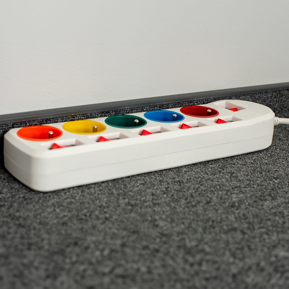 Biała listwa zasilająca z kolorowymi gniazdami, przy białej ścianie na szarej wykładzinie.