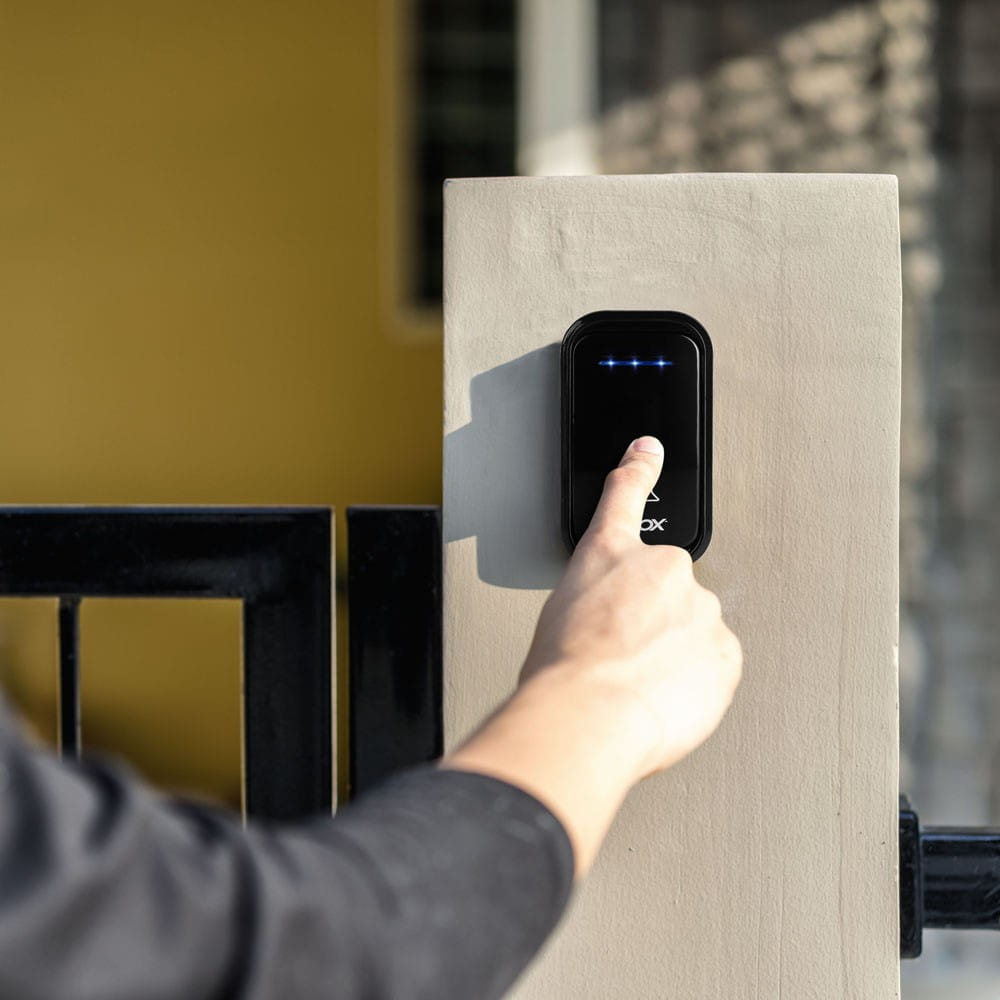 Ręka naciska czarny dzwonek na jasnoszarym filarze. Żółty budynek w tle.