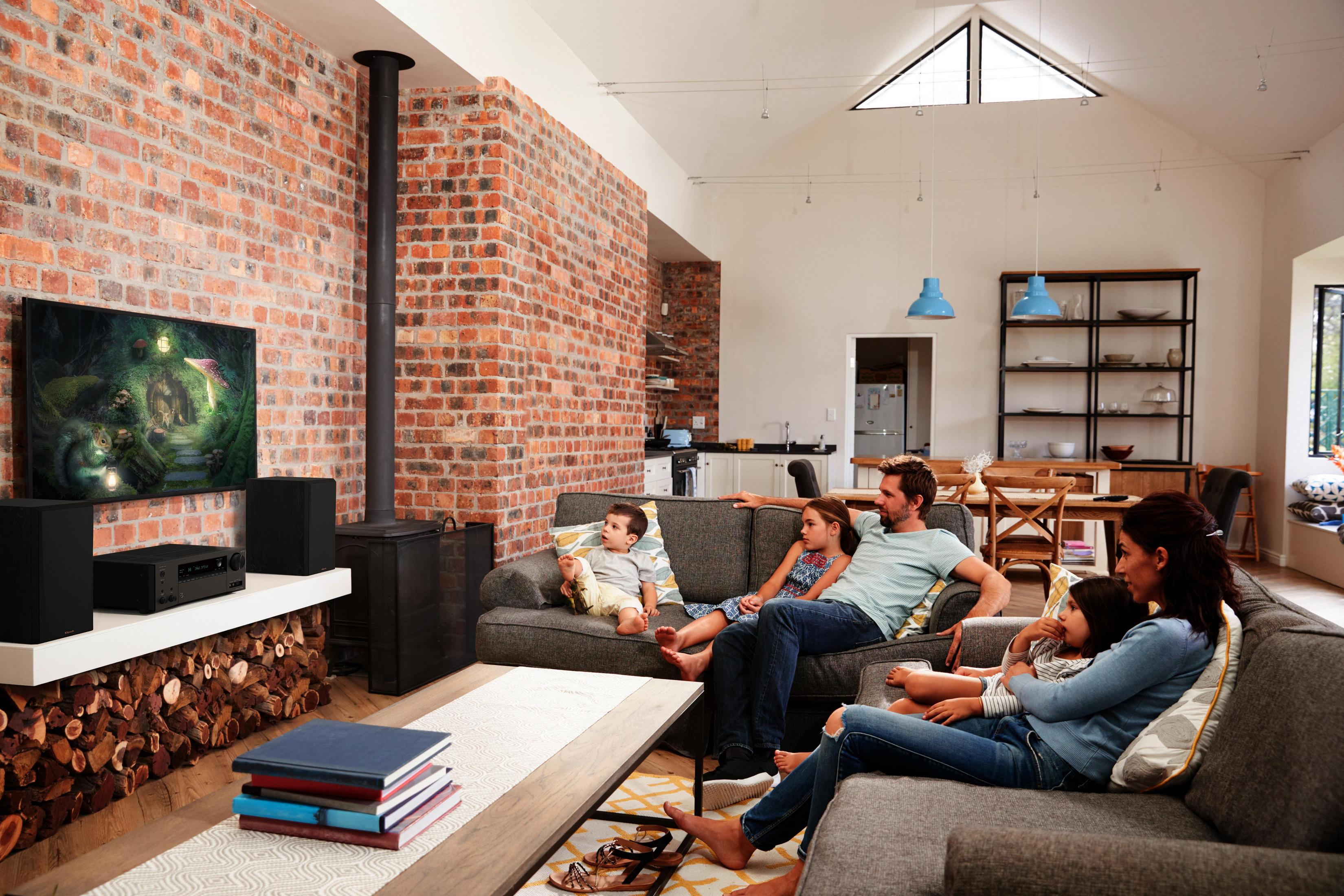 Rodzina ogląda telewizję w salonie. Widoczna ceglana ściana, kominek i półka z książkami.