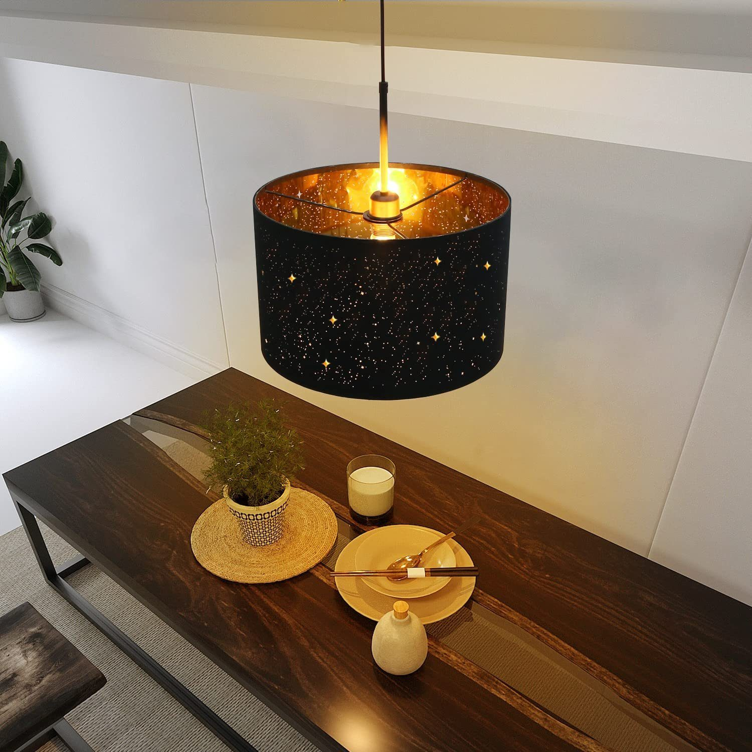 Draufsicht auf eine schwarze Pendelleuchte über einem Holztisch mit Gedecken. Eine kleine Pflanze steht auf dem Tisch.