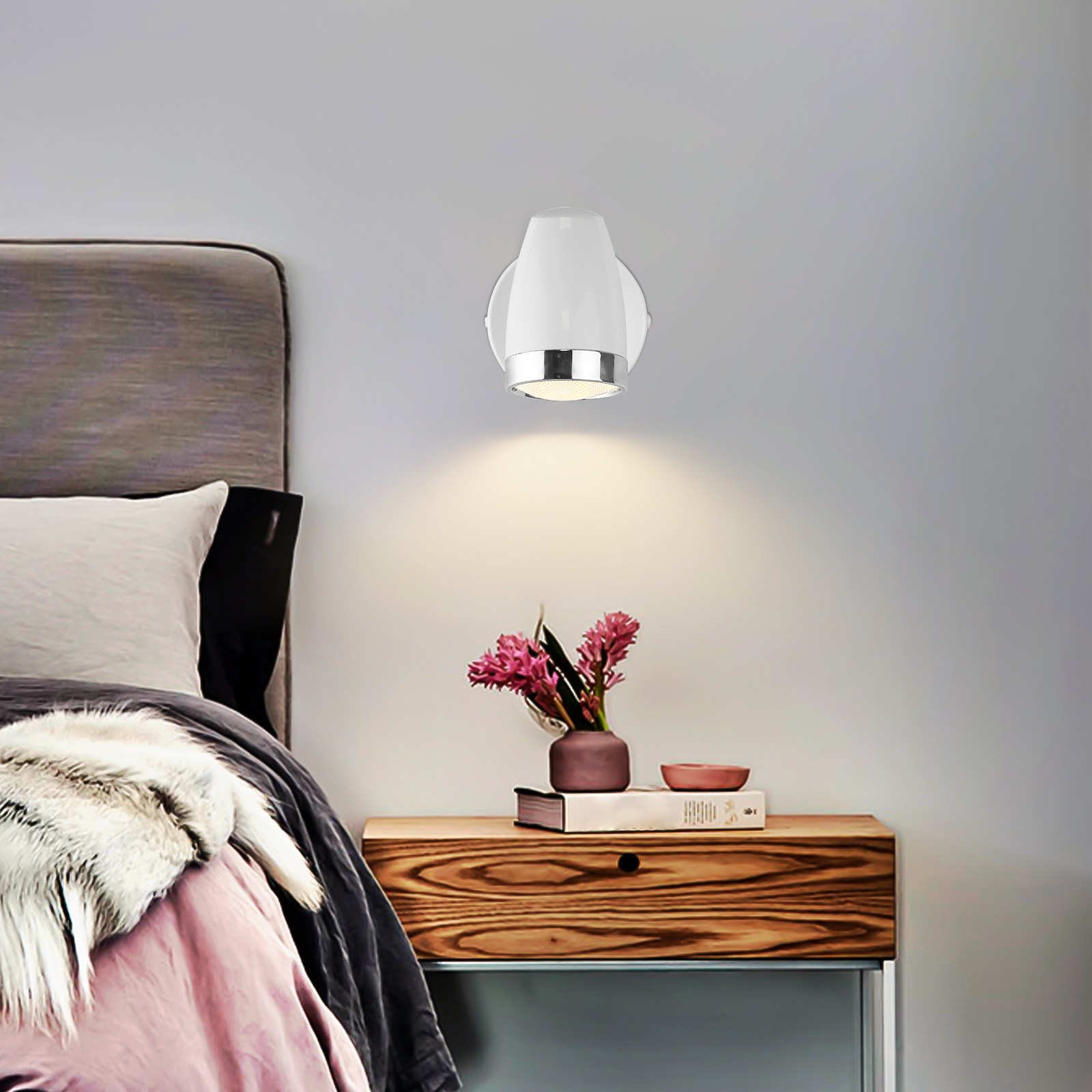 Biała lampa ścienna nad drewnianą szafką nocną. Różowe kwiaty i książka na szafce.