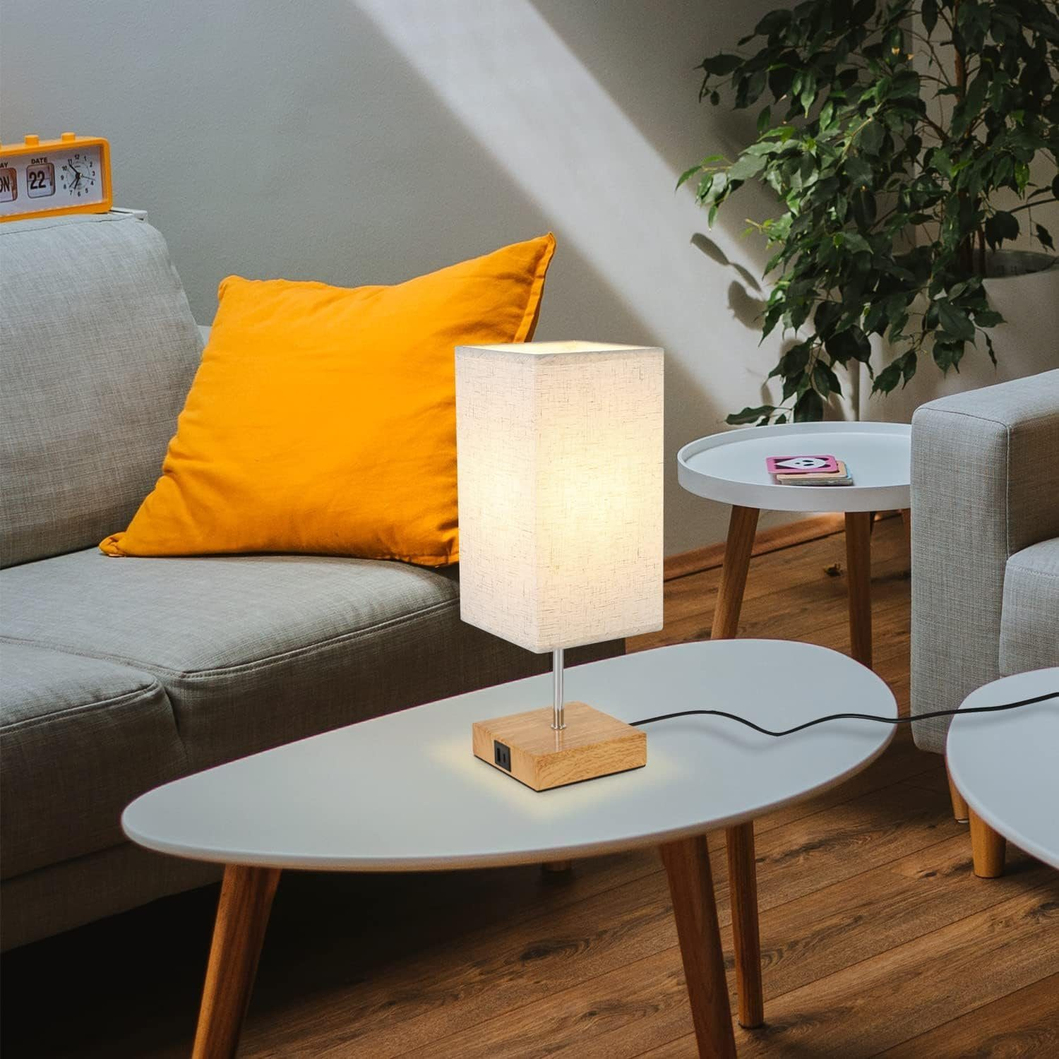 Oświetlona prostokątna lampa na białym stole. Żółta poduszka i zielona roślina w tle.