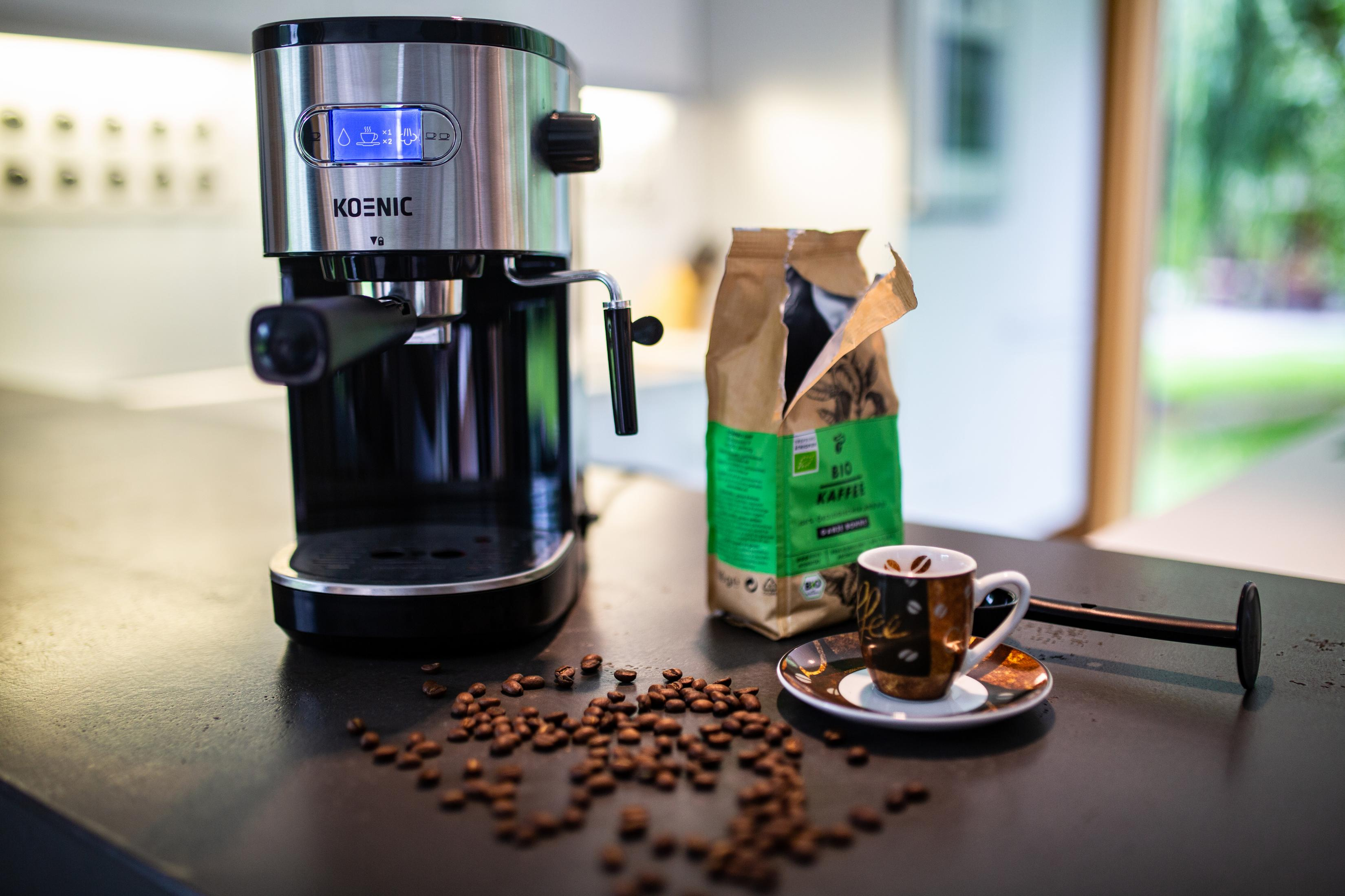 Una máquina de espresso junto a una bolsa de granos de café, una taza y un platillo, y granos derramados en una encimera.