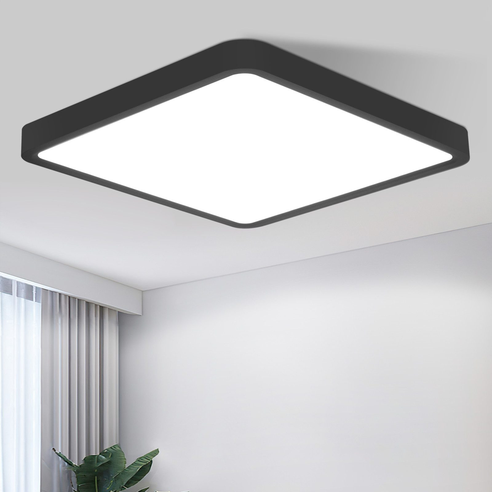 Eine quadratische schwarze Deckenlampe mit weißem Licht, vor einer weißen Wand. Es gibt eine grüne Pflanze.